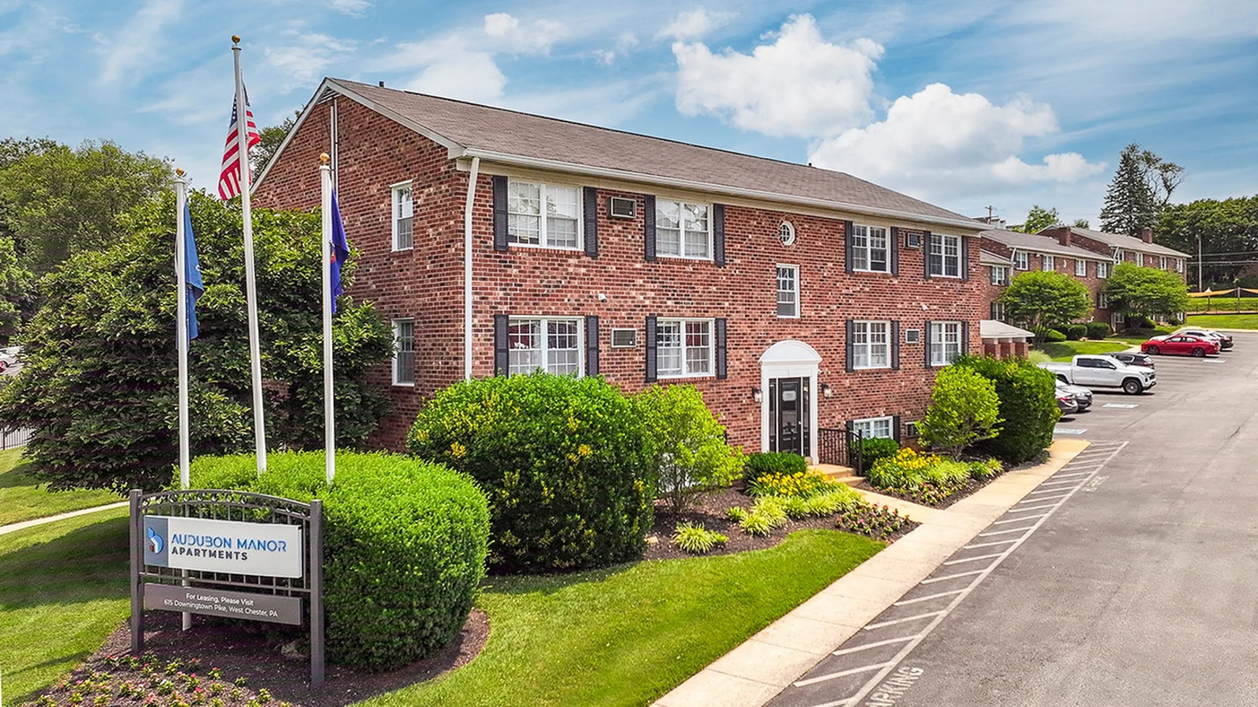 Charming brick apartment building surrounded by lush greenery, featuring a welcoming entrance and ample parking at Audubon Manor Apartments.