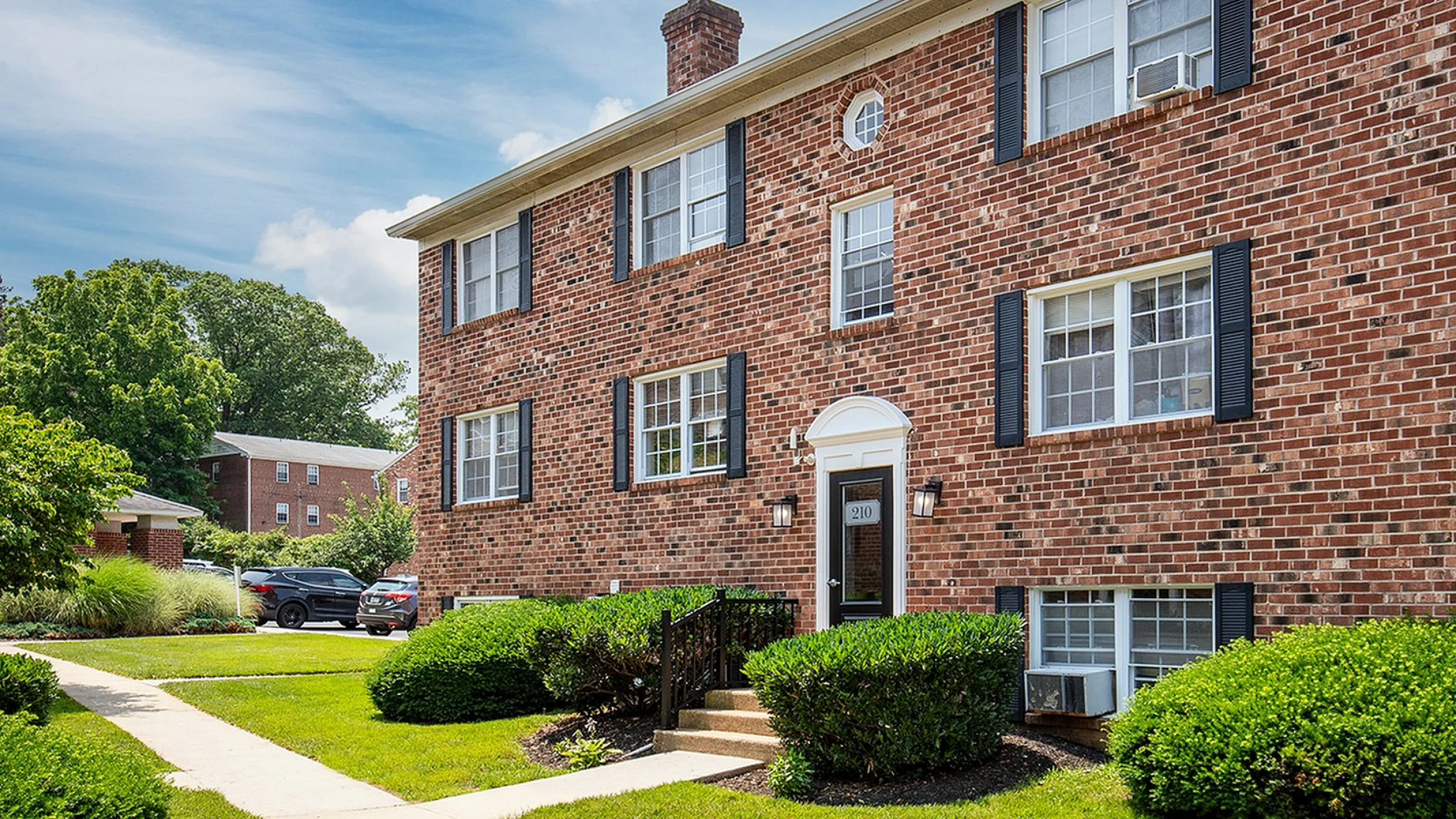 Charming brick apartment building with classic black shutters, well-maintained landscaping, and inviting walkway under a bright blue sky.