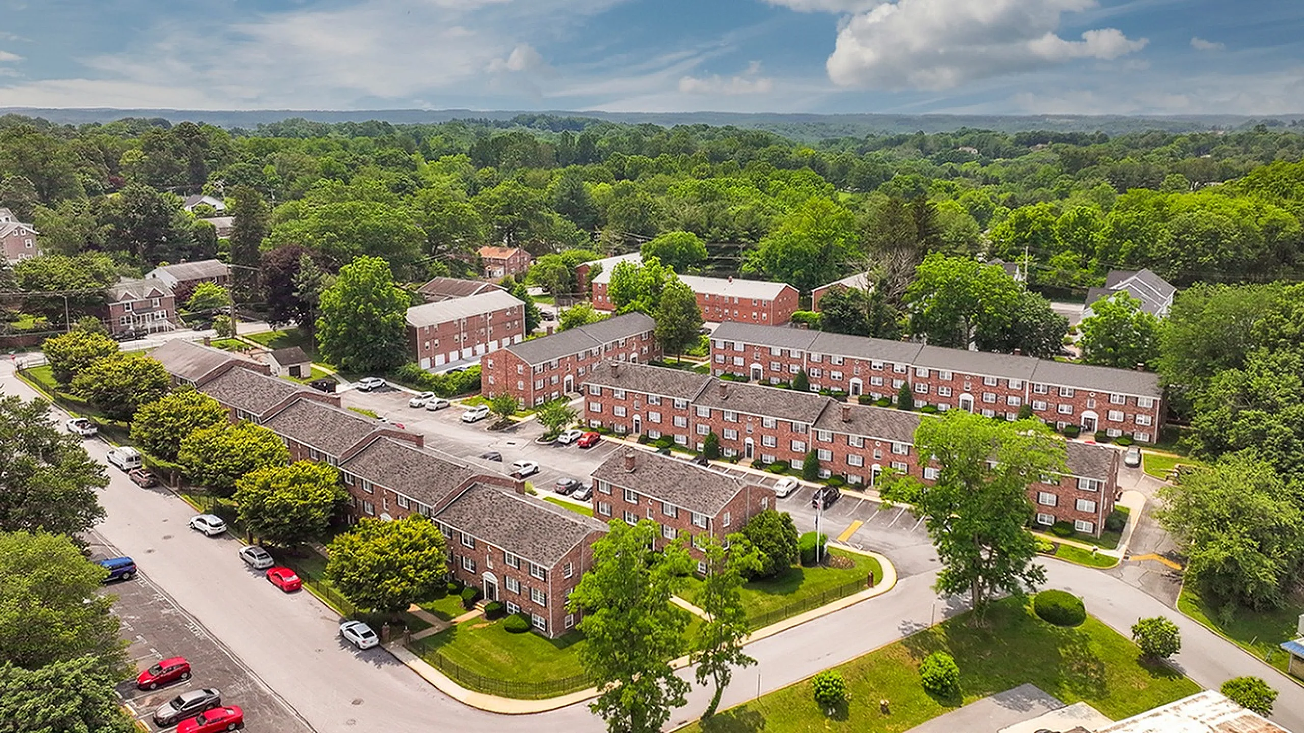 Charming aerial view of a serene apartment complex surrounded by lush greenery, featuring well-maintained brick buildings and ample parking space, perfect for comfortable living.