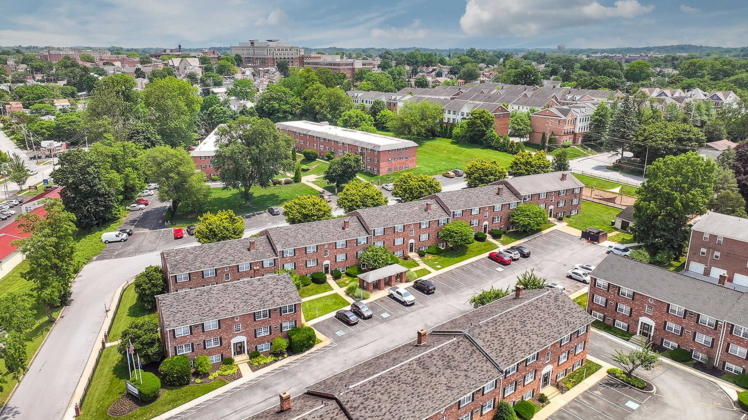 Aerial view of charming brick apartment complex surrounded by lush greenery, featuring ample parking and a vibrant neighborhood backdrop.
