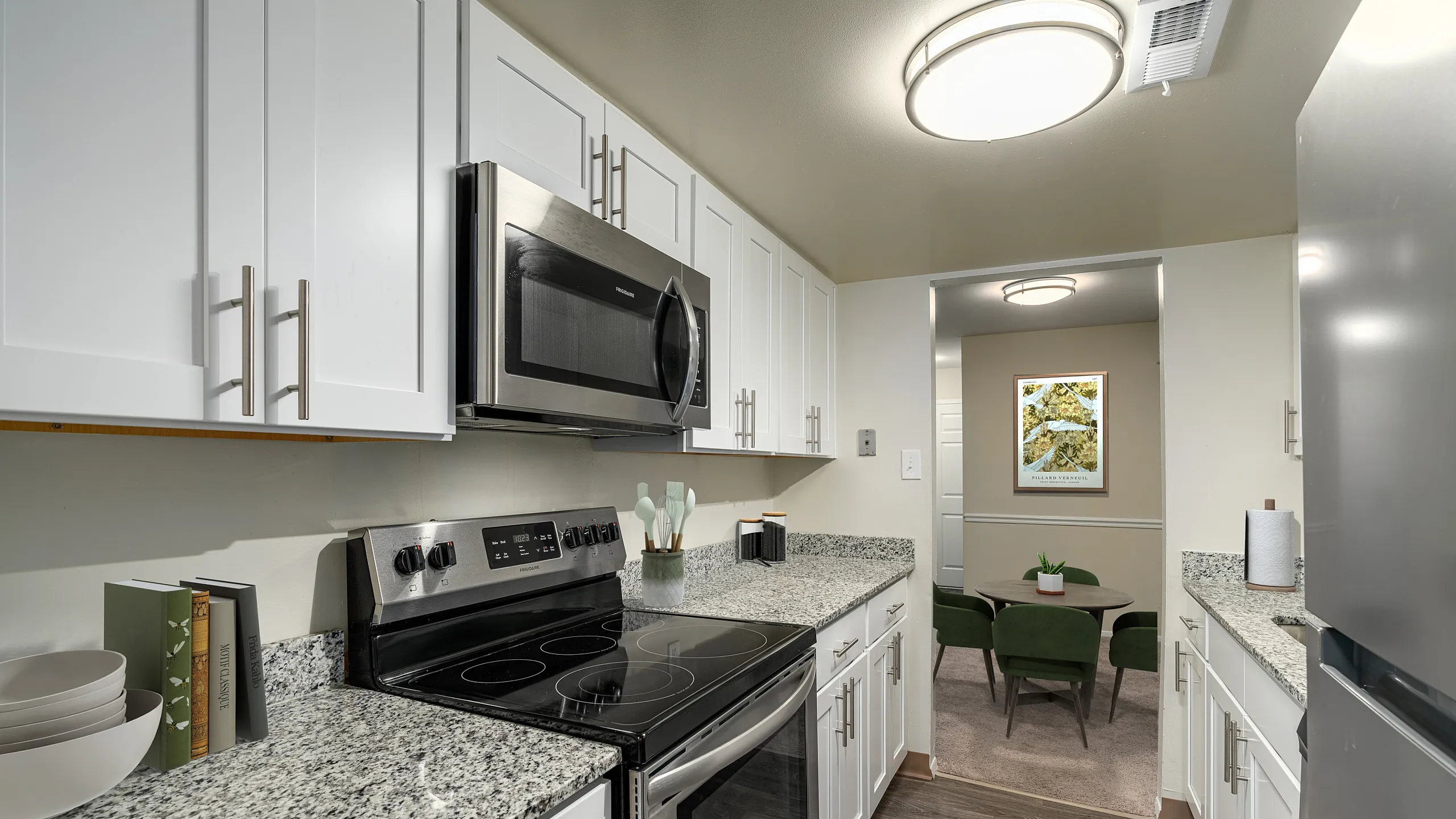 Modern kitchen with sleek granite countertops, stainless steel appliances, and stylish white cabinetry, adjacent to a cozy dining area with green velvet chairs.