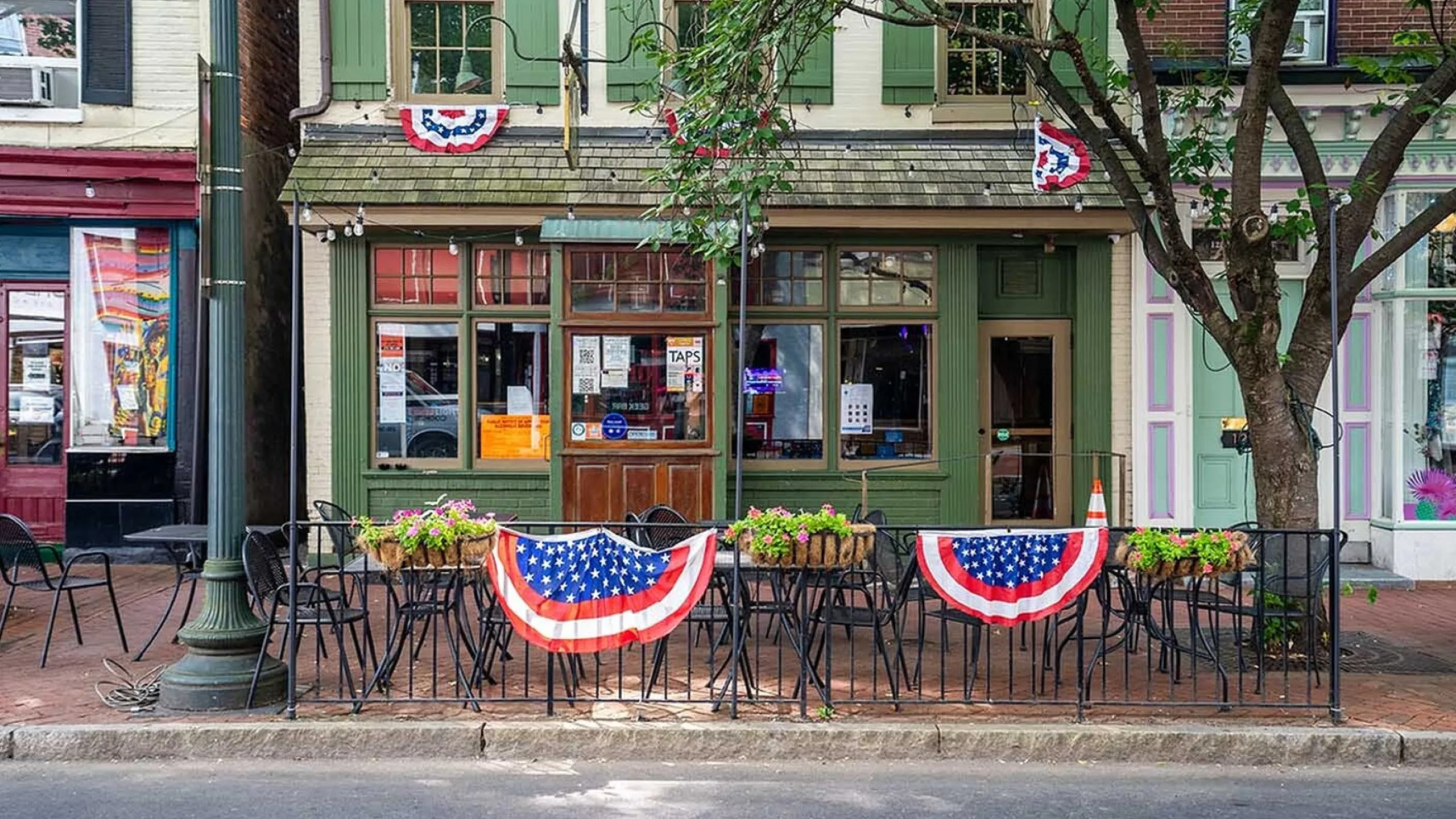 Charming street view of a cafe with patriotic American flags, outdoor seating, and vibrant flowers, inviting for a cozy dining experience.