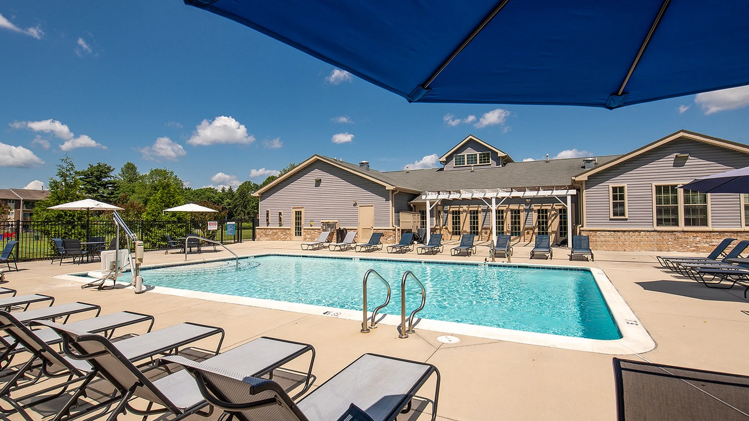 Inviting outdoor pool area with comfortable lounge chairs, surrounded by lush greenery and a modern clubhouse under a clear blue sky.