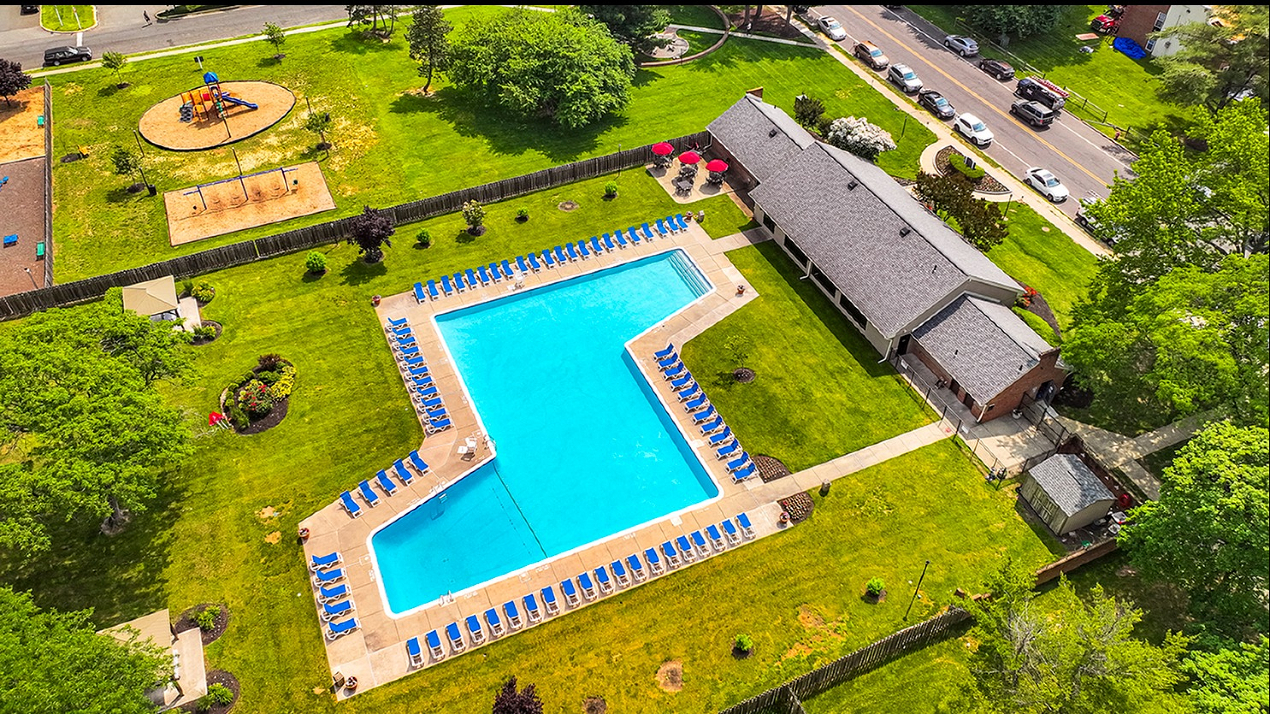 Aerial view of a luxurious community pool area with numerous blue sun loungers, surrounded by lush green lawns and landscaped gardens, adjacent to a charming clubhouse and a family-friendly playground.