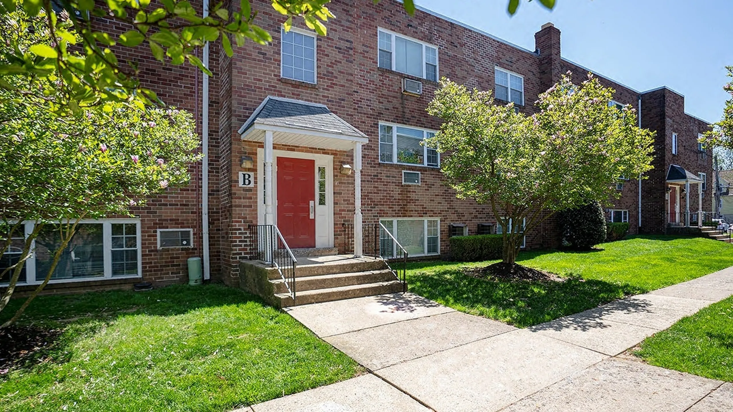 Charming red-brick apartment building with lush green landscaping and inviting red doors, perfect for comfortable living.