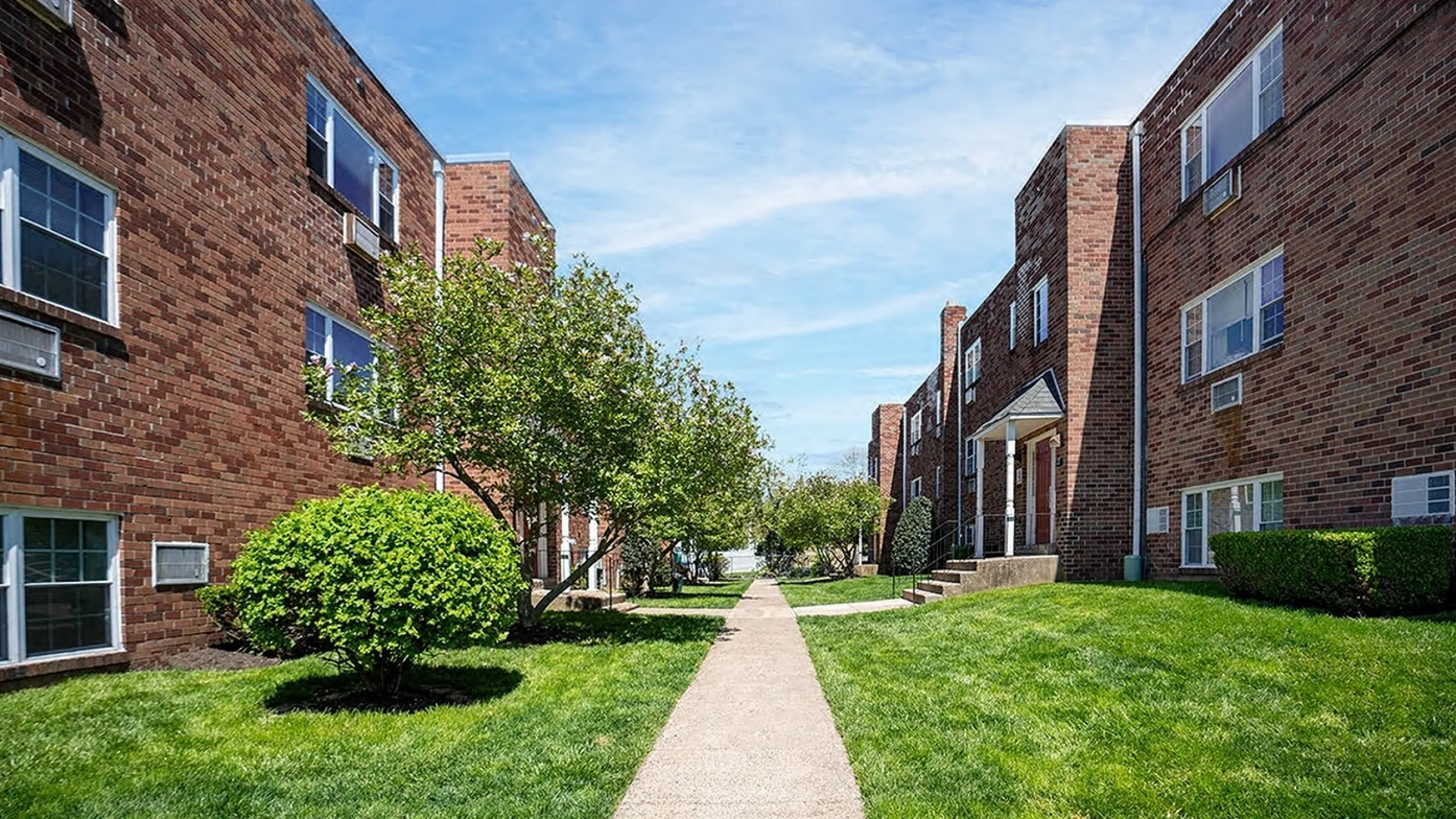 Charming brick apartment complex with lush green landscaping and a central pathway under a bright blue sky.