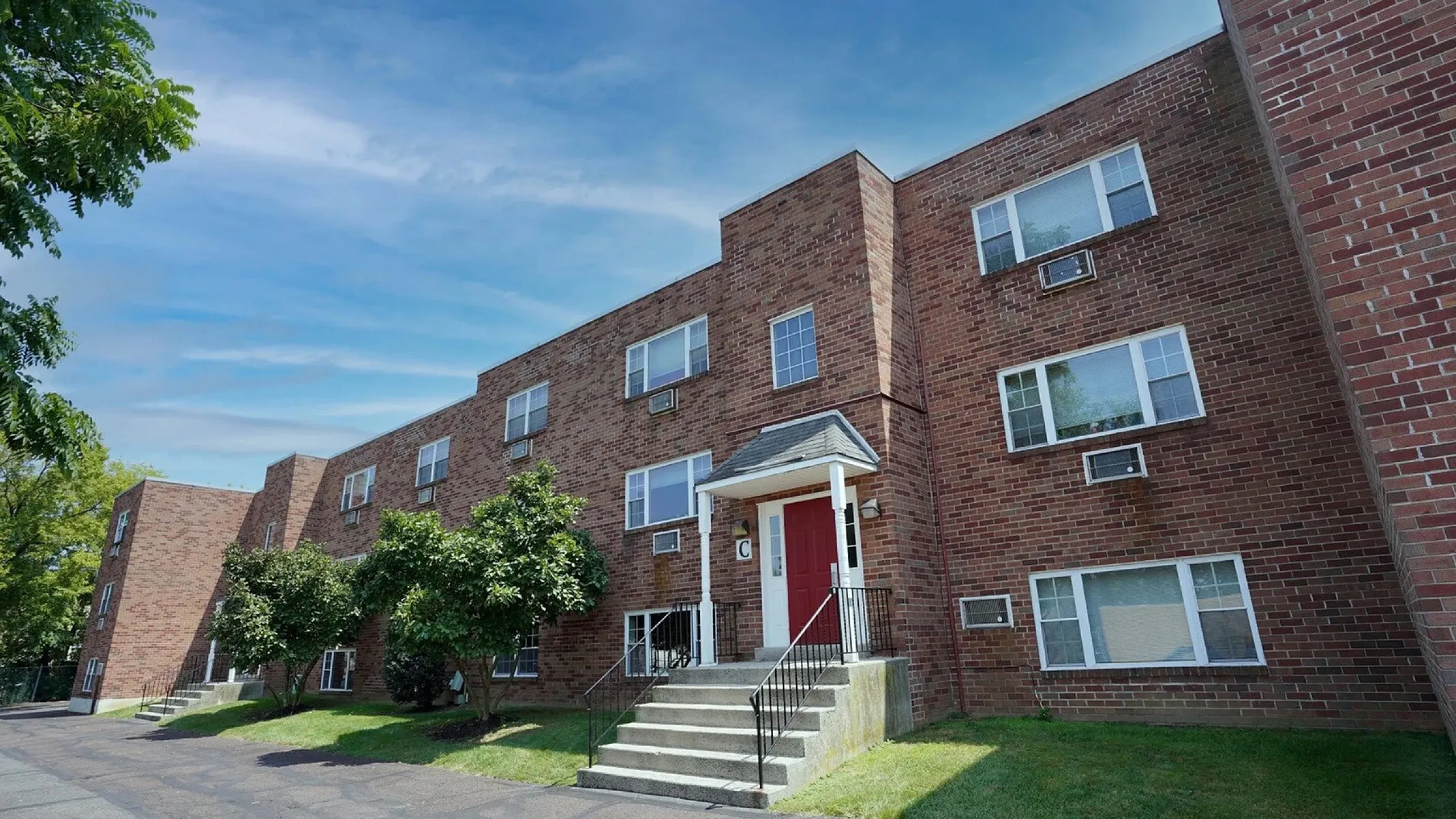 Charming brick apartment building with lush green surroundings and a welcoming entrance under a bright blue sky.