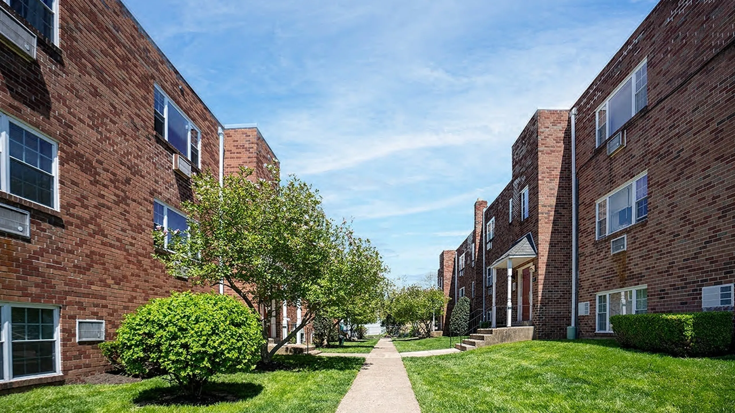 Charming red brick apartment buildings with lush green lawns and trees, bathed in bright sunshine, offering a welcoming and serene living environment.