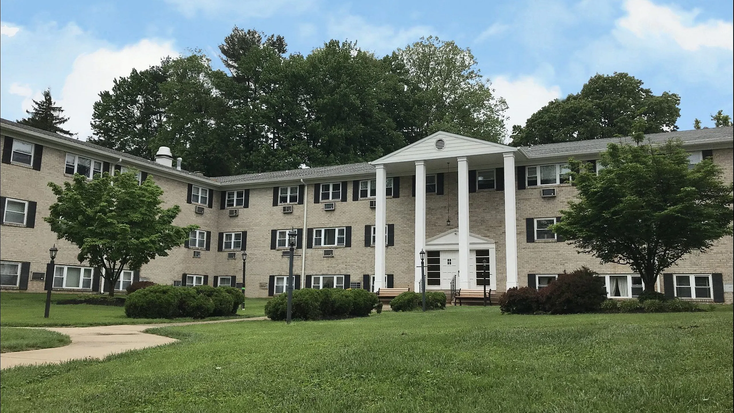 Charming brick apartment building surrounded by lush greenery, featuring a classic entrance with white columns, perfect for tranquil living.