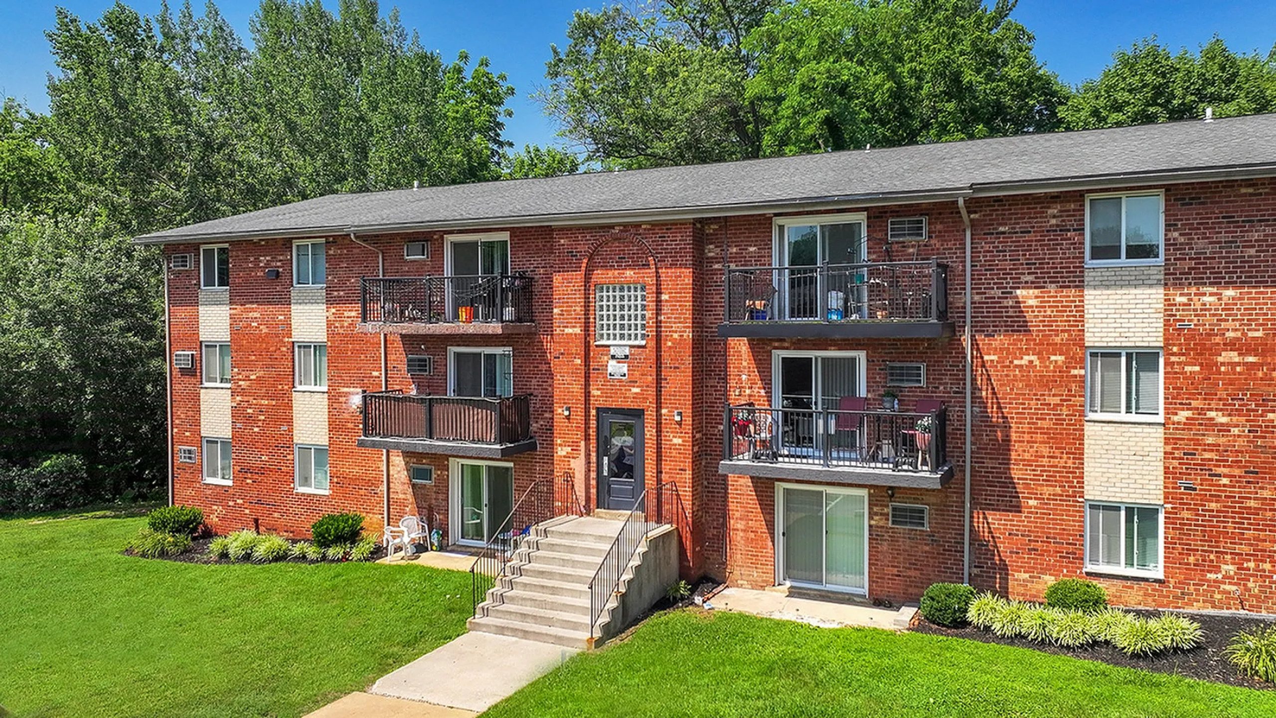 Charming red brick apartment building with elegant balconies, surrounded by lush greenery, offering a welcoming and serene living space.