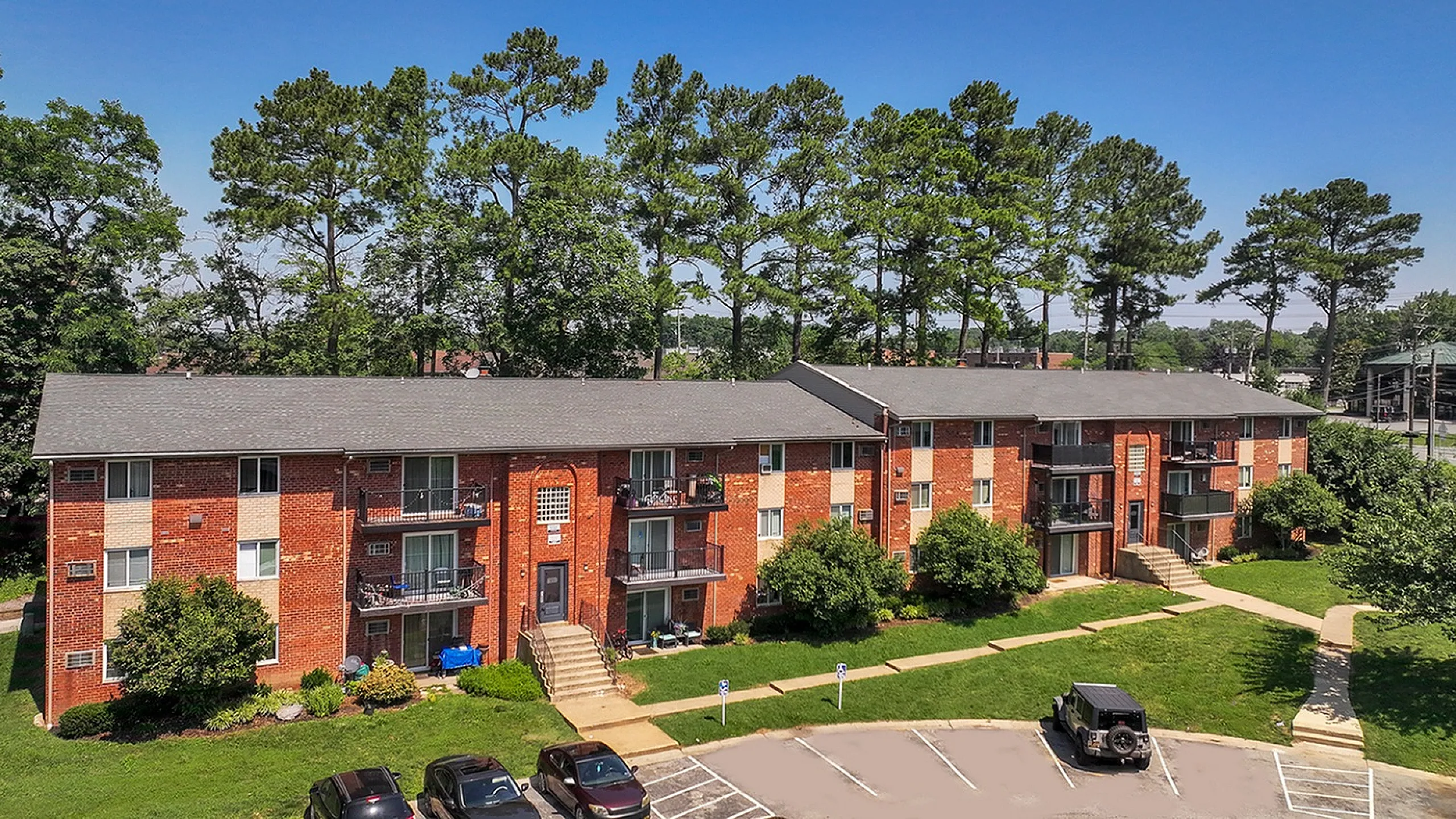 Charming brick apartment complex surrounded by lush greenery and tall trees, featuring multiple balconies and ample parking space.