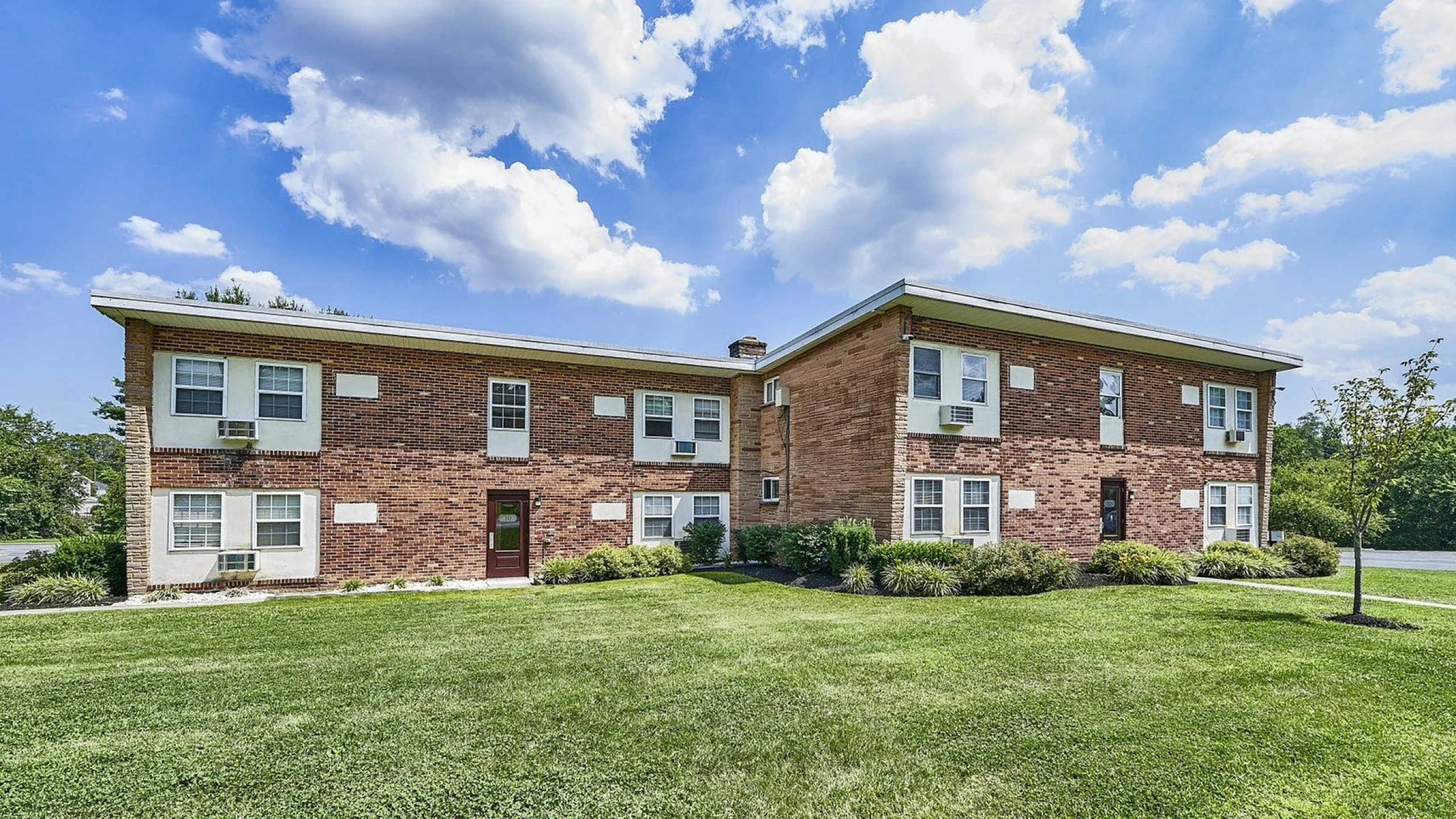 Charming brick apartment building with lush green lawn, surrounded by blue skies and picturesque clouds.