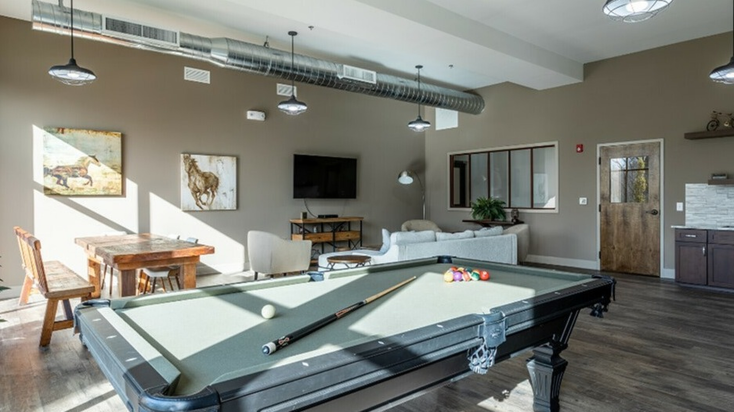 Luxury apartment lounge with a modern pool table, cozy seating area, and stylish decor. Sunlit ambiance with a rustic dining table, artwork, and open kitchenette.