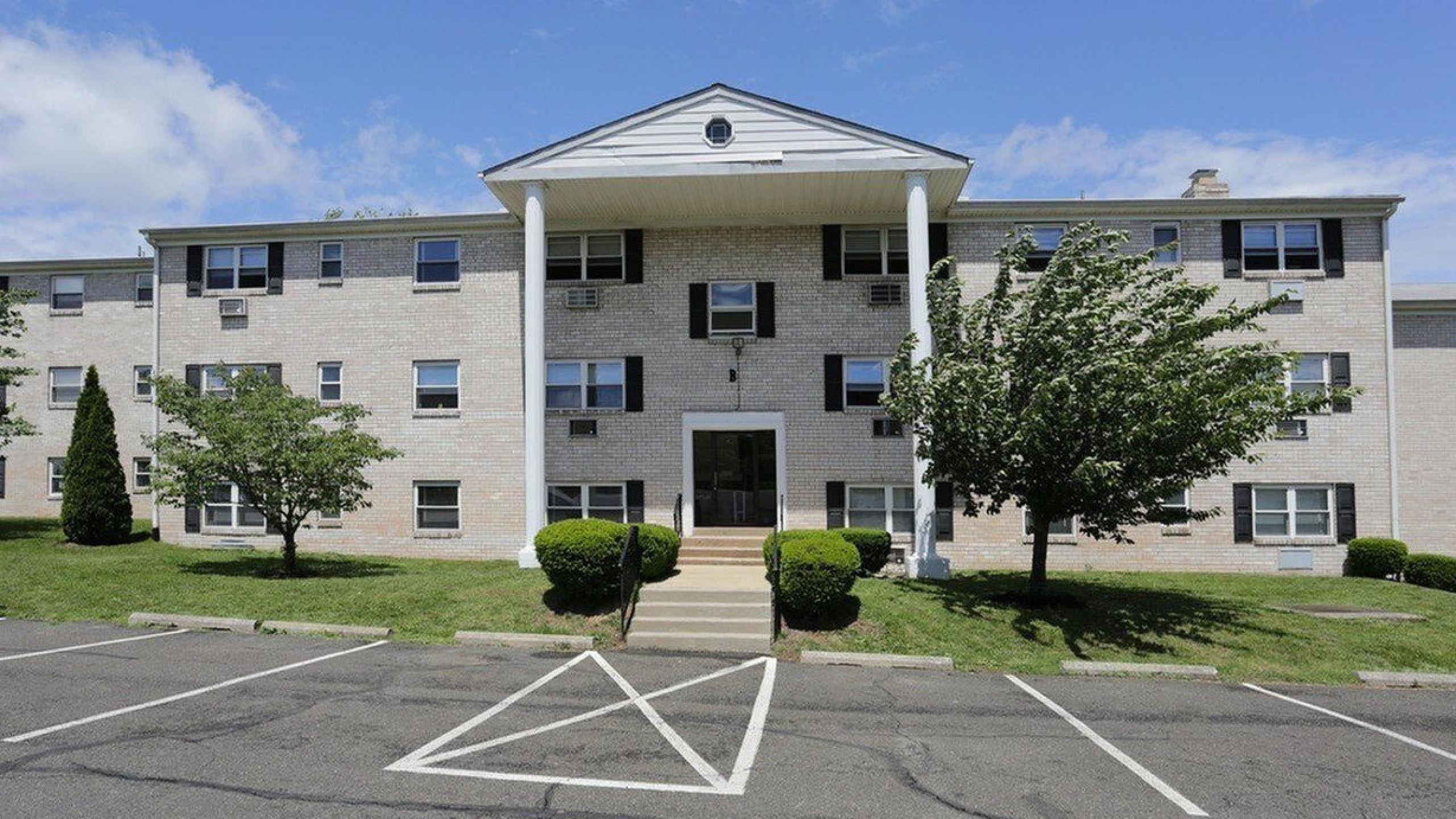Charming apartment building with classic colonial design, lush green landscaping, and ample parking space under a clear blue sky.