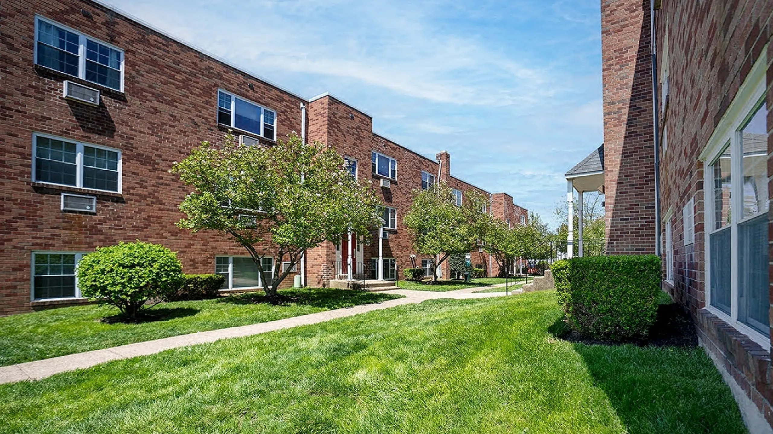 Charming brick apartment complex surrounded by lush greenery and well-maintained pathways under a clear blue sky.