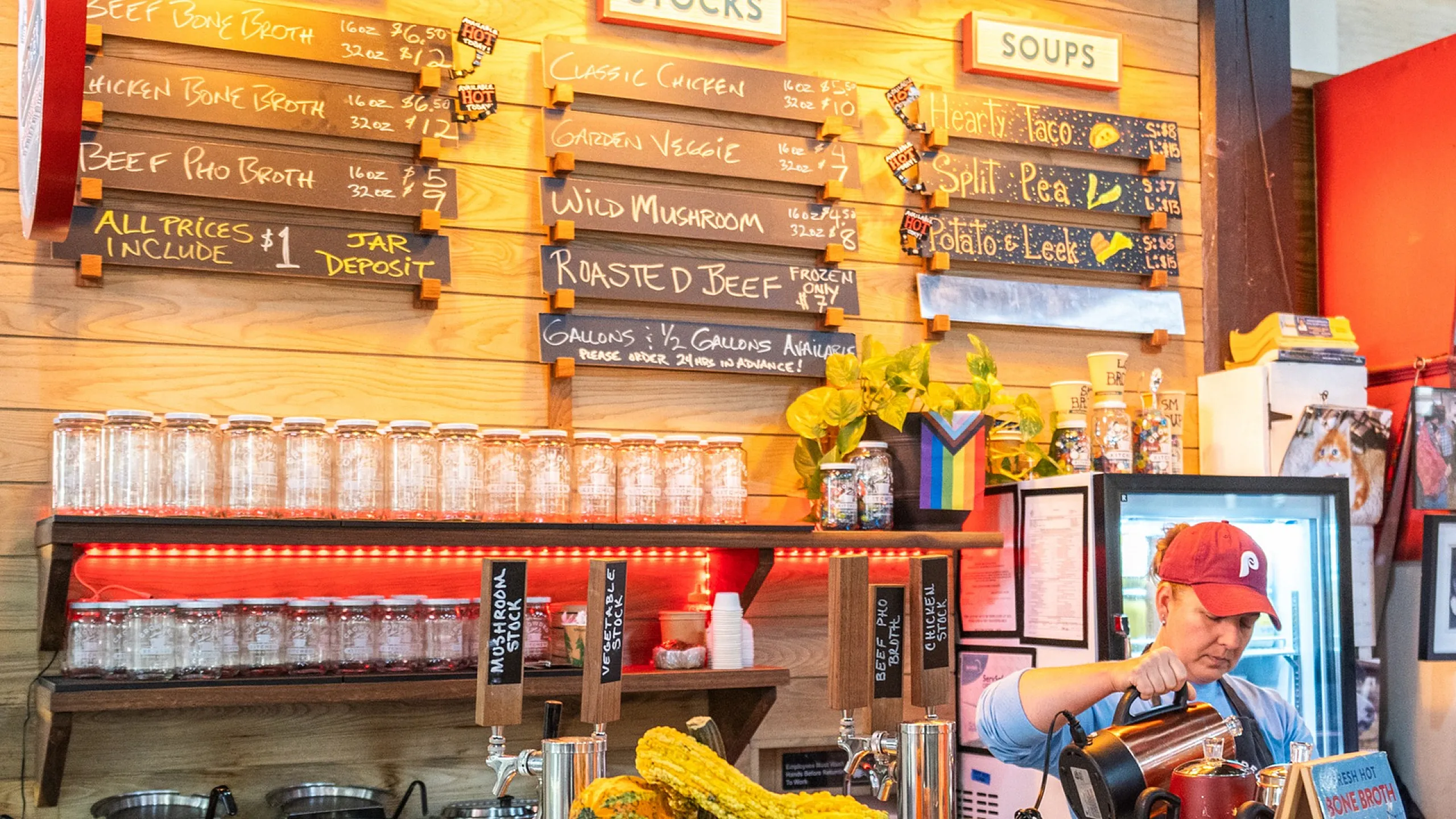 Vibrant soup counter with a variety of broths and soups, displayed alongside a cheerful server preparing fresh ingredients beneath warm lighting.