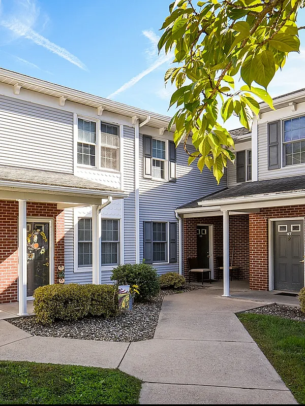 Charming suburban townhouse with brick and siding exterior, surrounded by lush greenery under a bright blue sky.