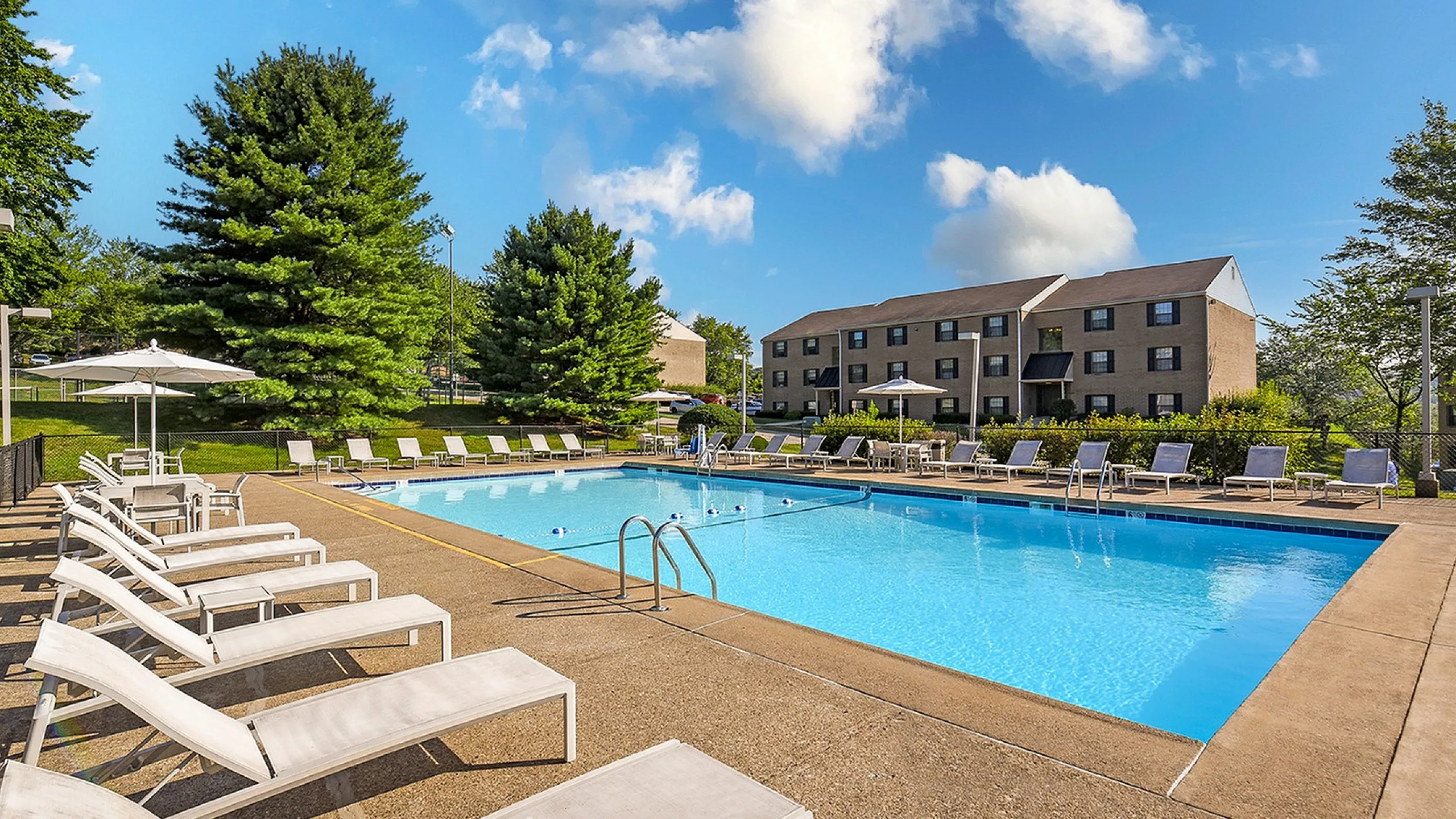 Inviting outdoor pool with sun loungers and umbrellas, surrounded by lush greenery and modern apartment buildings under a clear blue sky.
