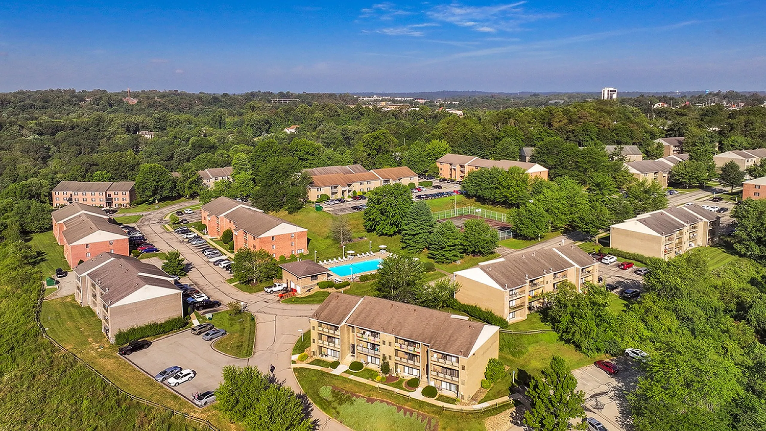 Charming aerial view of a vibrant apartment community nestled in lush greenery, showcasing modern buildings, a sparkling swimming pool, and ample parking.