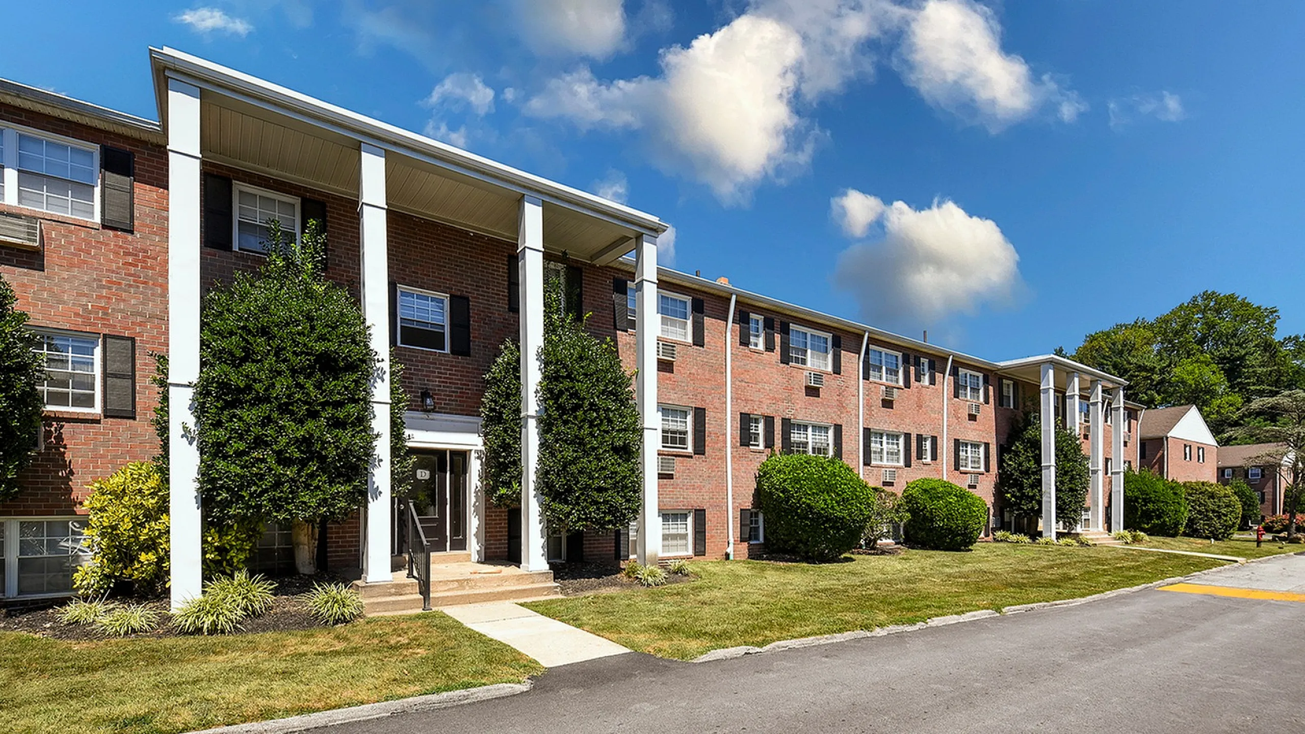 Charming apartment building with a classic brick facade, surrounded by lush greenery and set against a bright blue sky, offering a serene and inviting living environment.