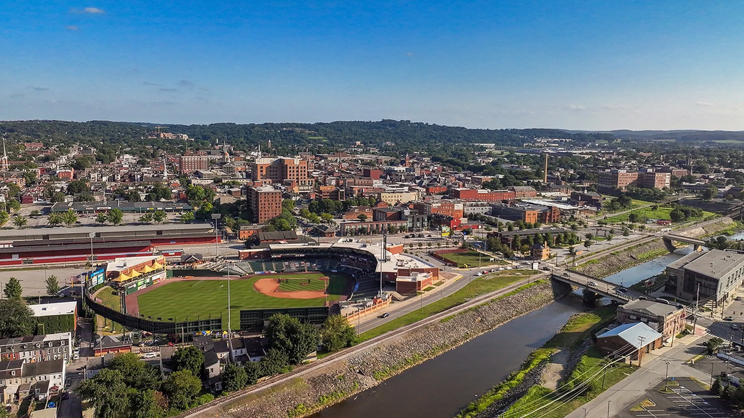 Vibrant aerial view of a charming cityscape featuring a picturesque baseball stadium, surrounded by lush greenery and historic architecture, with a serene river meandering through the town.