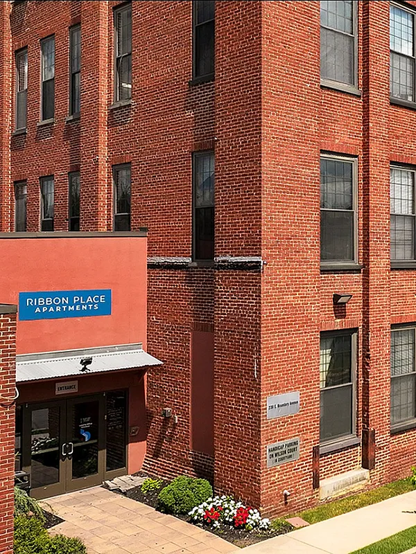 Charming red-brick exterior of Ribbon Place Apartments with lush landscaping and a welcoming entrance, perfect blend of historic charm and modern living.