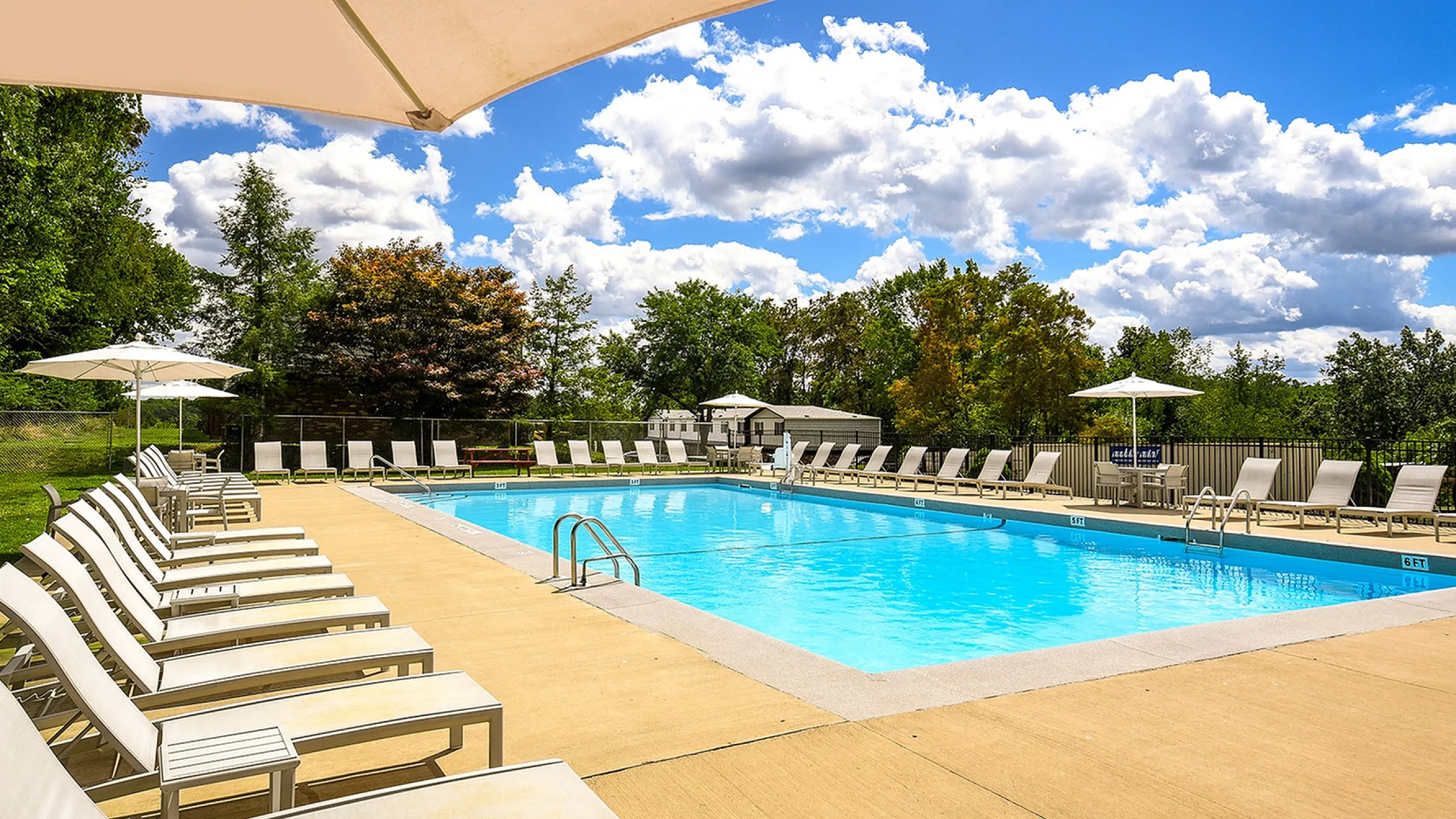 Inviting outdoor swimming pool surrounded by elegant lounge chairs and vibrant greenery under a bright blue sky with fluffy clouds.