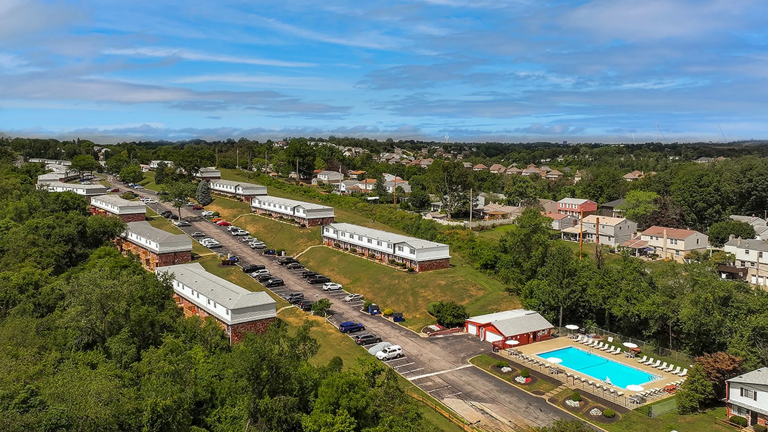 Charming hillside apartment complex with well-maintained buildings, ample parking, and a sparkling blue swimming pool surrounded by lush greenery.