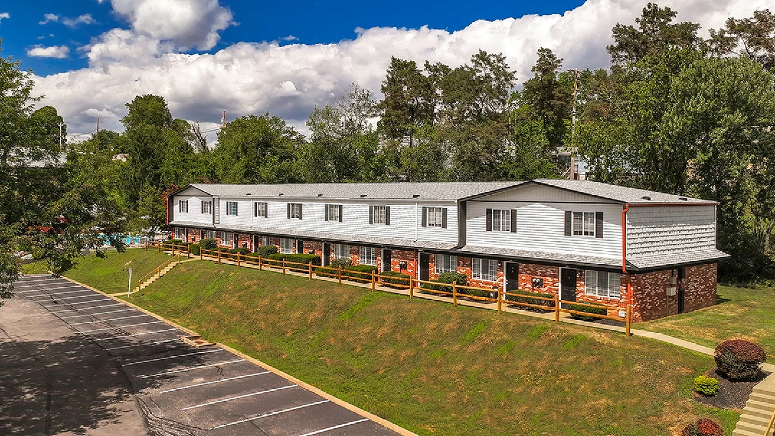 Charming townhouse complex surrounded by lush greenery, featuring ample parking and vibrant landscaping under a bright blue sky.