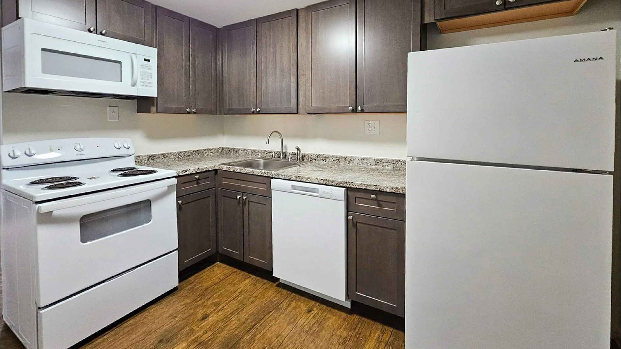 Modern kitchen with sleek dark cabinets, a white stove, microwave, dishwasher, and refrigerator, complemented by elegant granite countertops and warm hardwood flooring.