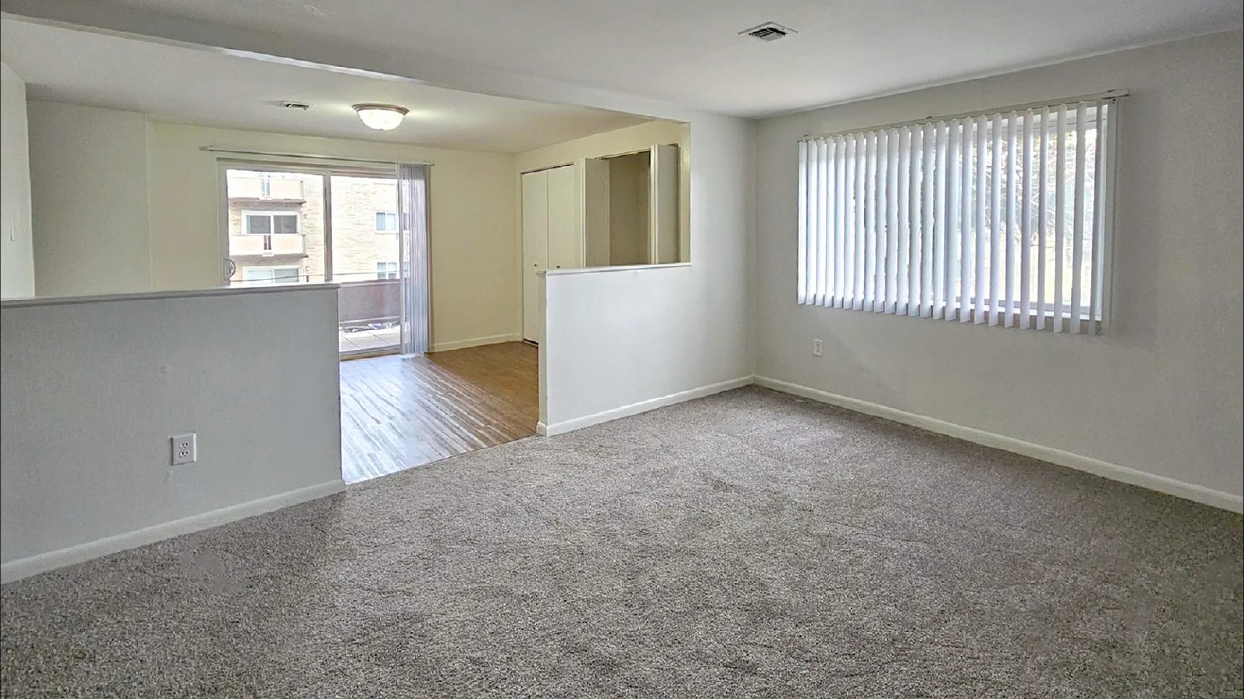 Bright and spacious living area with large windows, carpet flooring, and open flow to a hardwood space, ideal for seamless living.