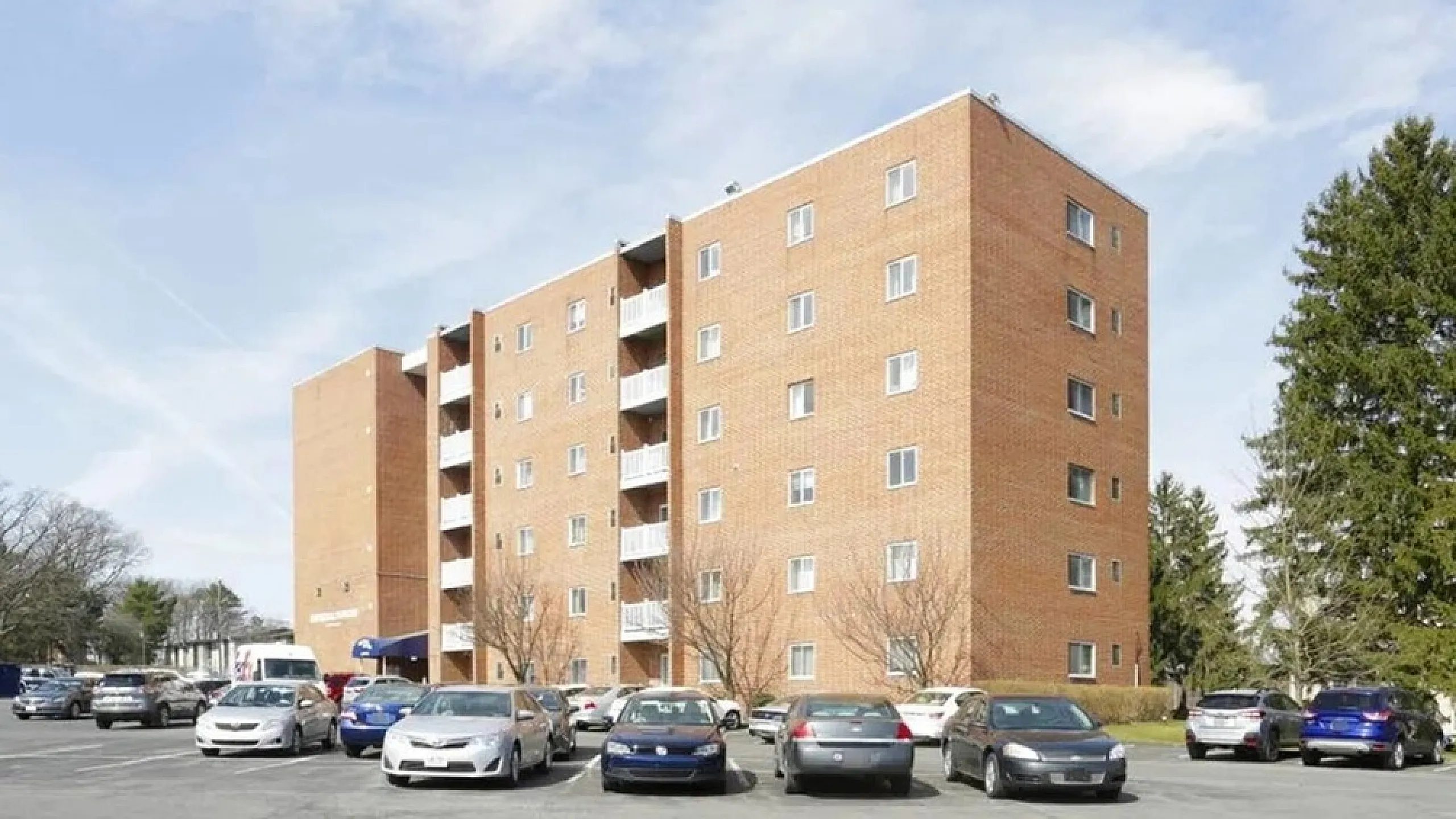 Charming brick apartment building with spacious parking area, surrounded by greenery and blue skies.
