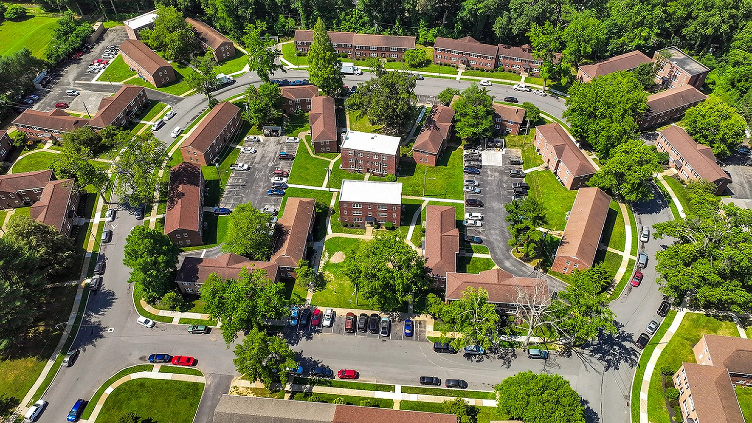 Vibrant aerial view of a charming apartment complex surrounded by lush greenery and ample parking spaces, offering a serene community living experience.