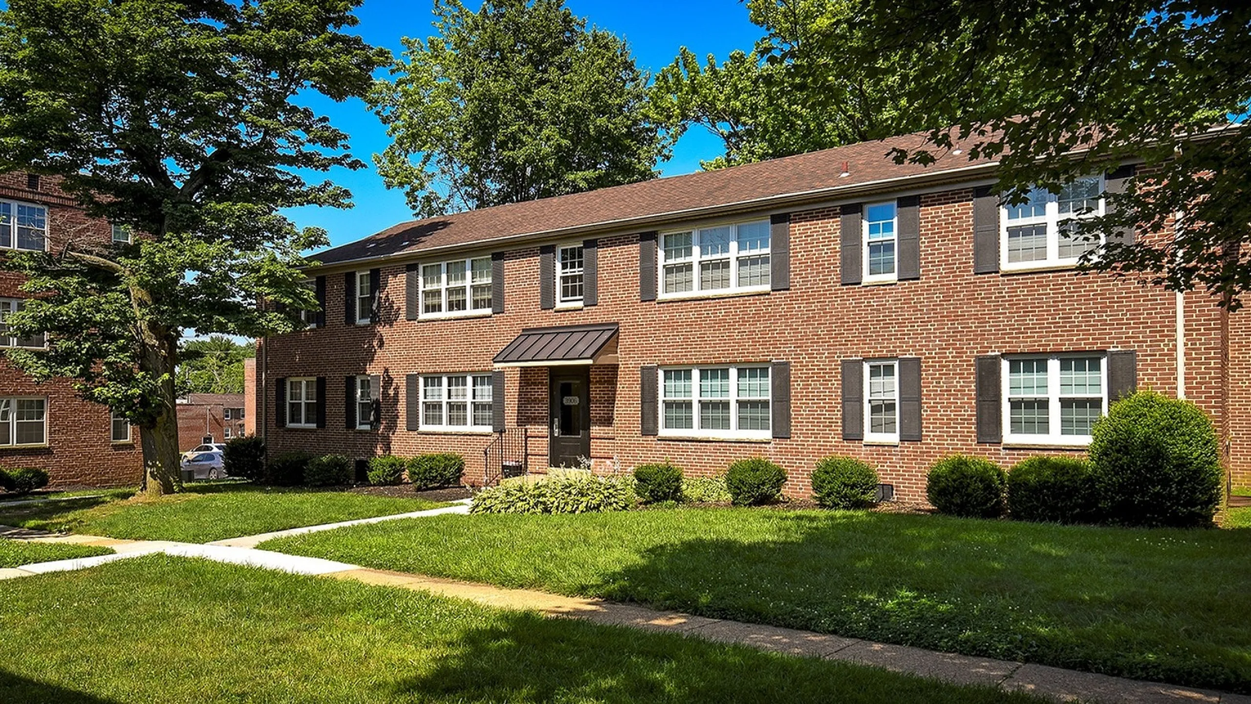 Charming brick apartment building with lush green landscaping and mature trees under a clear blue sky.