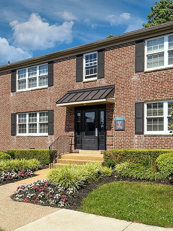Charming brick apartment building surrounded by lush greenery and vibrant flower beds under a clear blue sky.