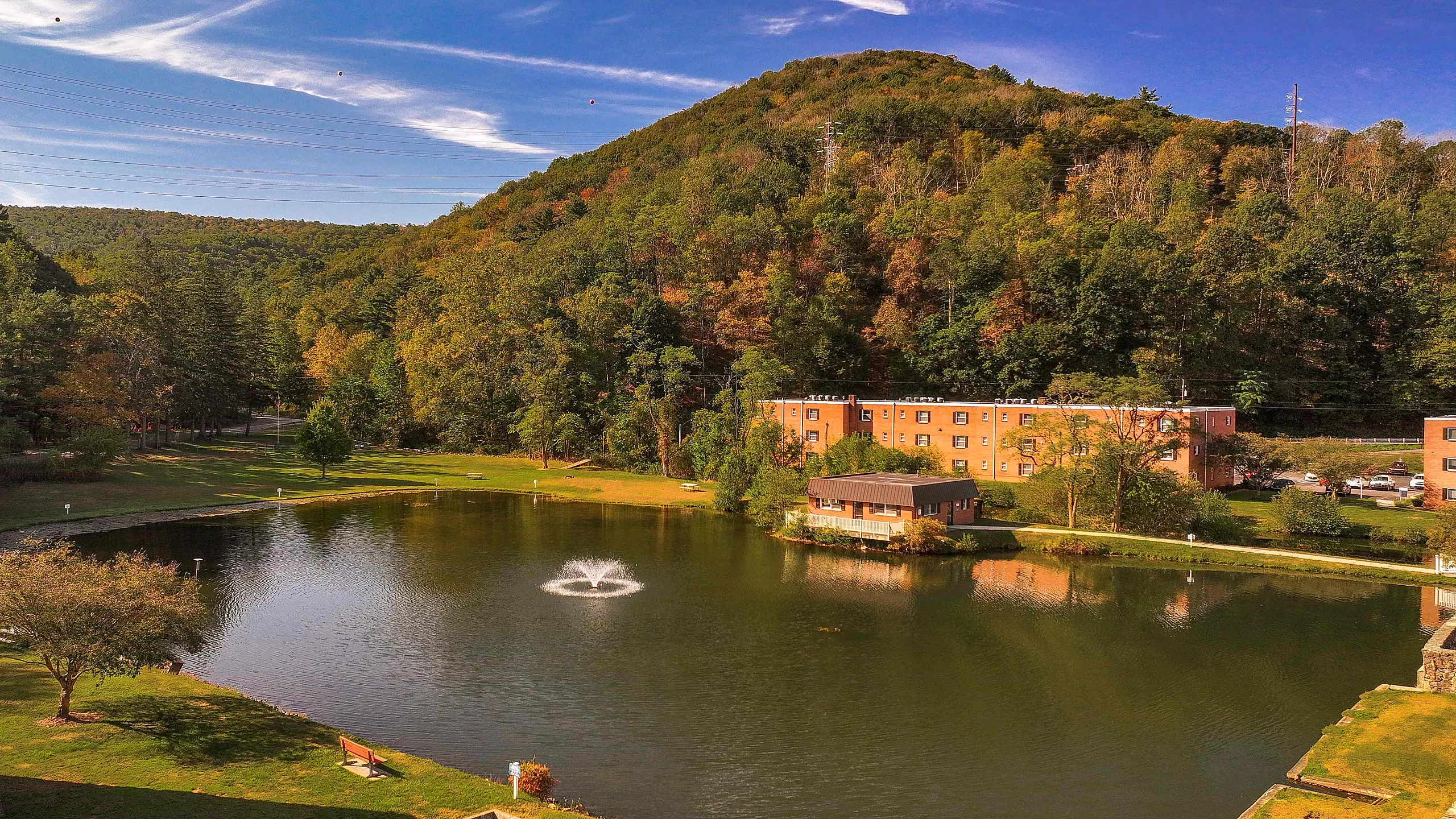 Scenic view of serene pond with a fountain, surrounded by lush trees and a charming apartment building nestled against a picturesque hillside.