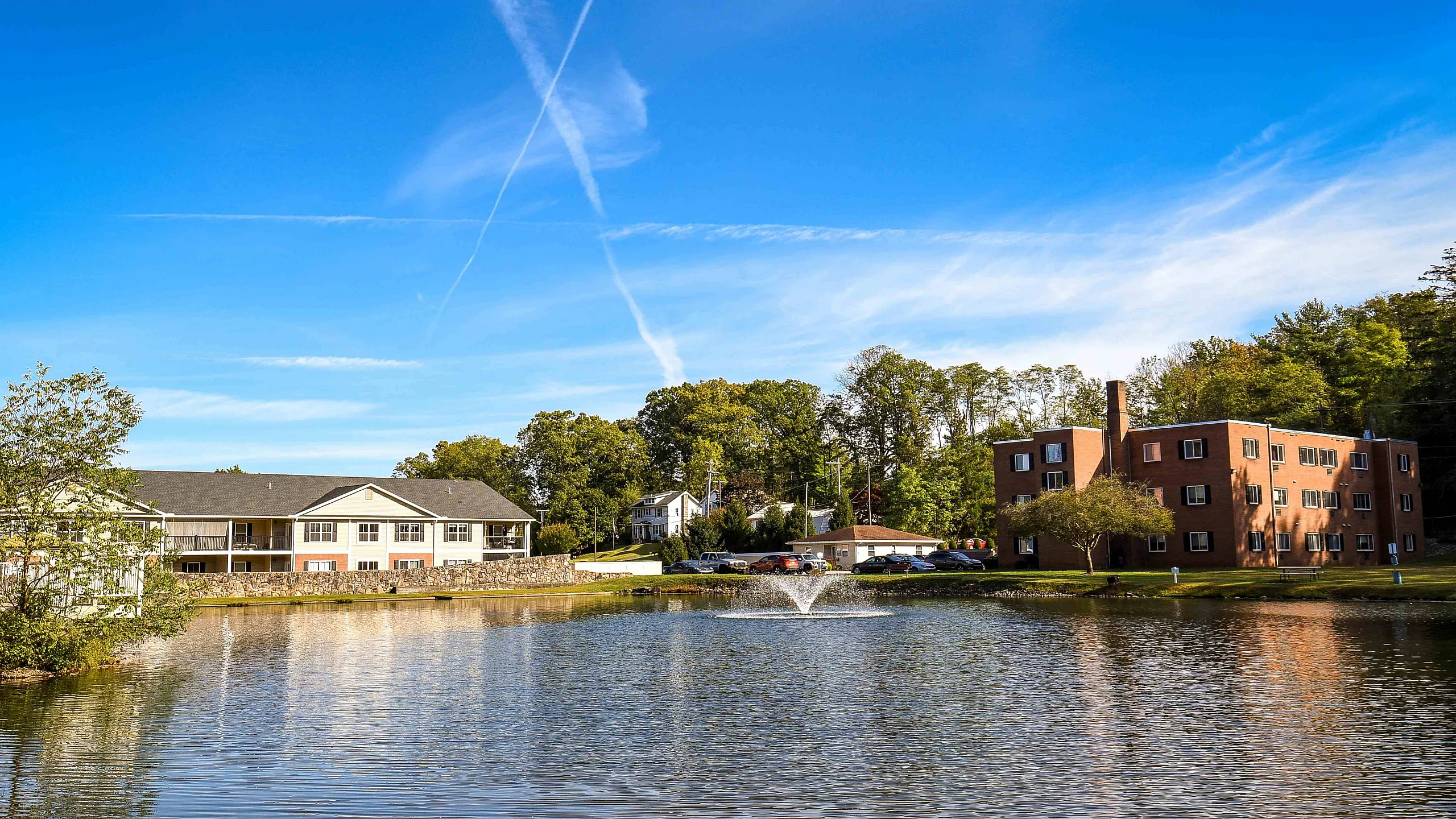 Serene apartment complex with scenic lake view, surrounded by lush trees and featuring charming brick buildings under a clear blue sky.