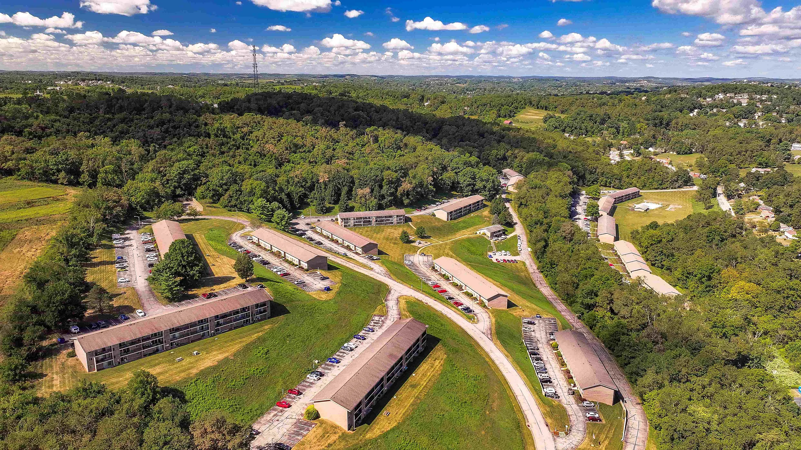 Scenic aerial view of apartment complex surrounded by lush greenery, showcasing numerous buildings with spacious parking and serene landscape.