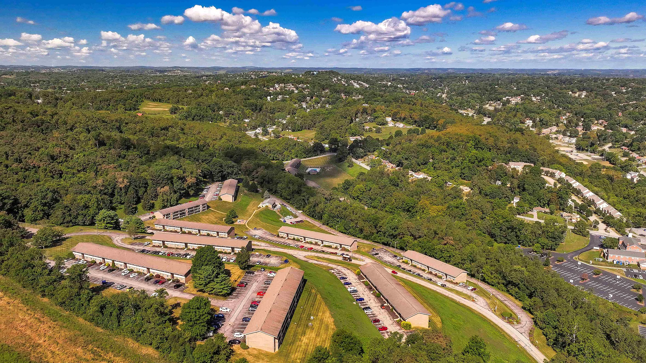 Scenic aerial view featuring a sprawling community nestled amidst lush greenery, highlighting residential buildings with ample parking and serene surroundings.