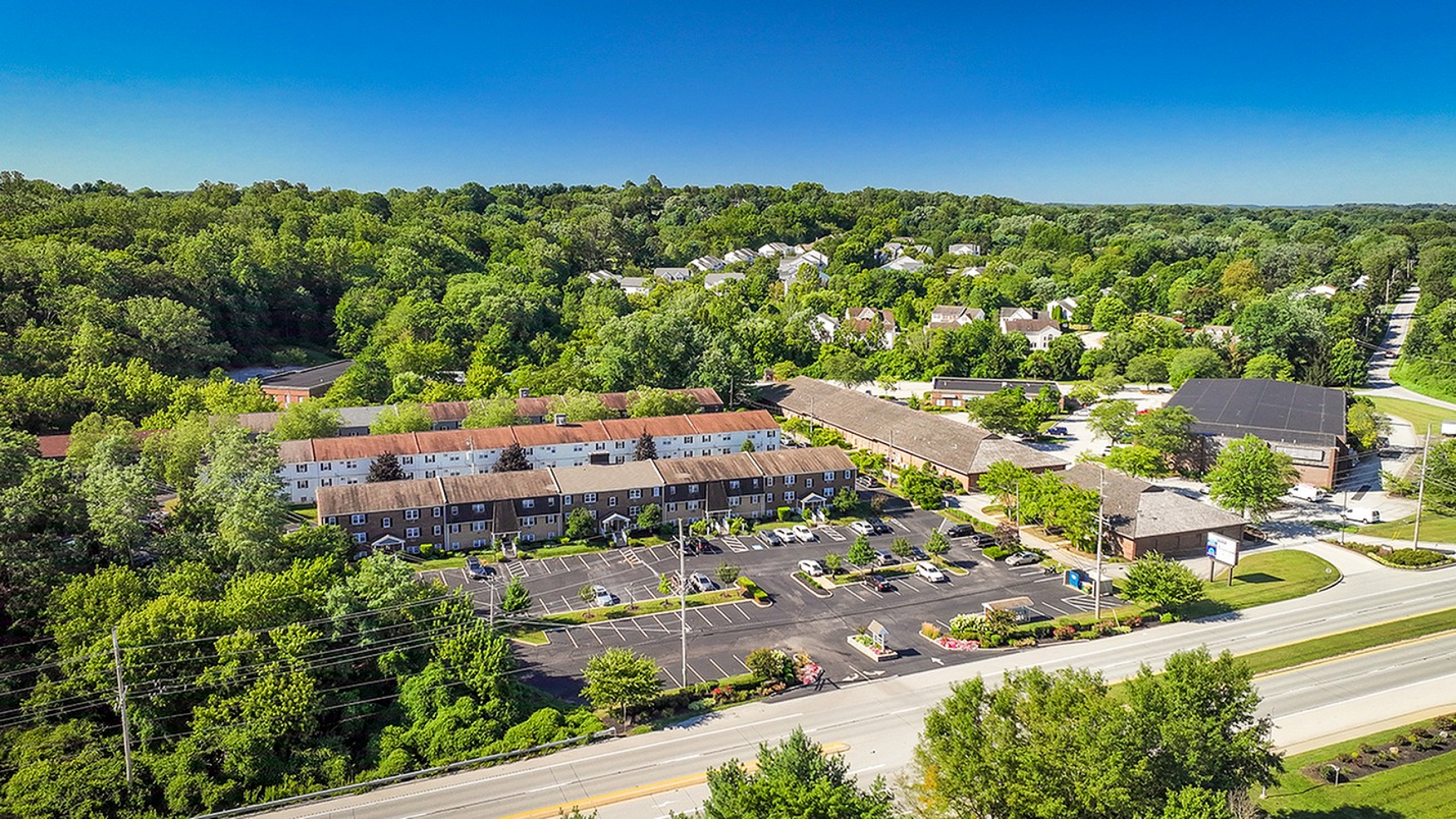 Aerial view of charming apartment complex surrounded by lush greenery and featuring ample parking, capturing a serene and inviting residential setting.