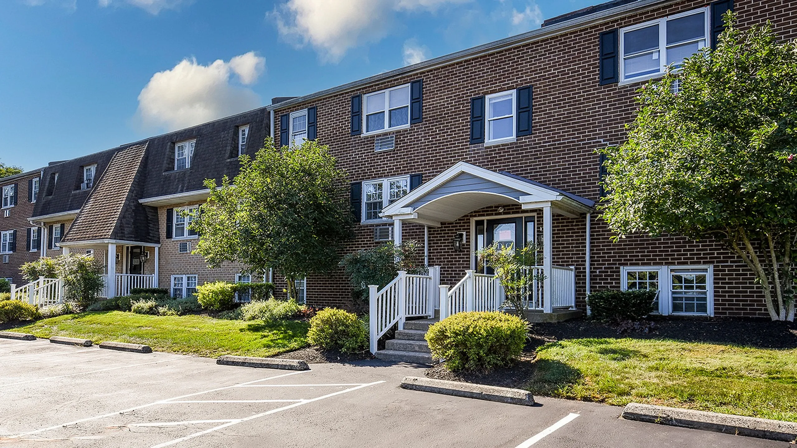 Charming brick apartment building with manicured landscaping under a bright blue sky, featuring welcoming entryway and ample parking.