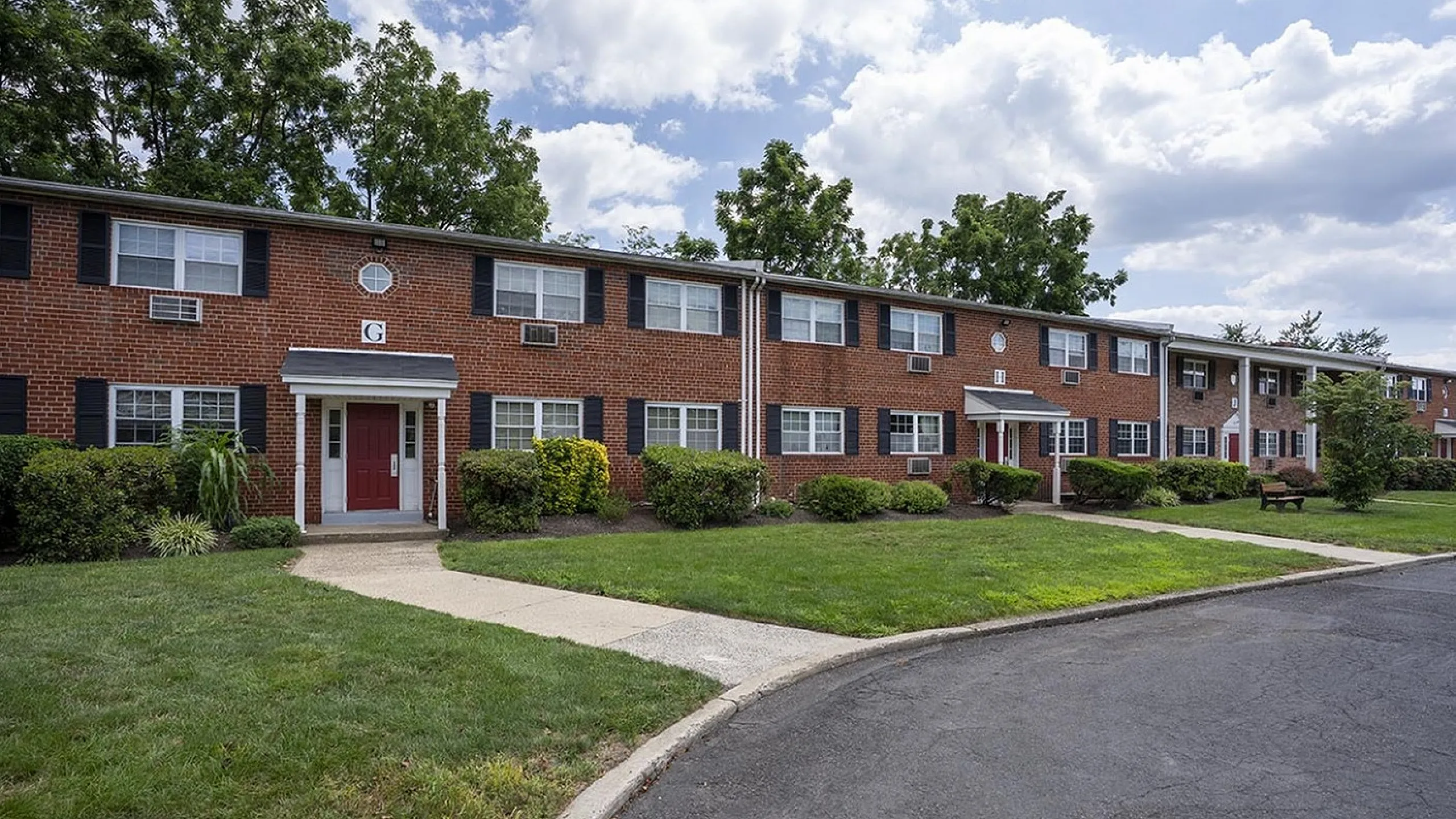 Charming brick apartment complex with lush green landscaping and inviting pathways, set against a clear blue sky backdrop.