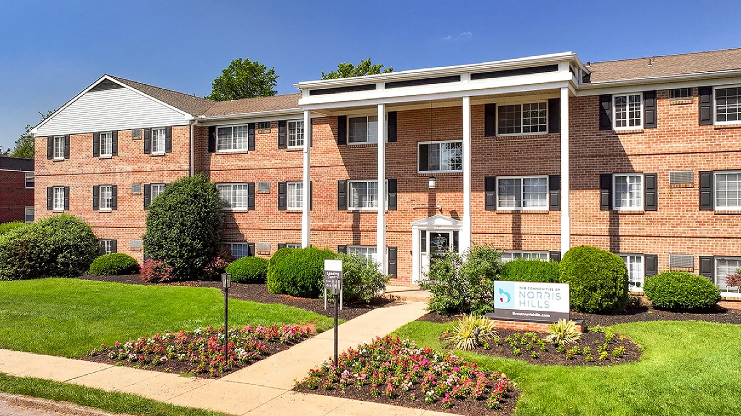 Charming brick apartment building with lush landscaping and vibrant flower beds, offering a welcoming atmosphere in a serene setting.