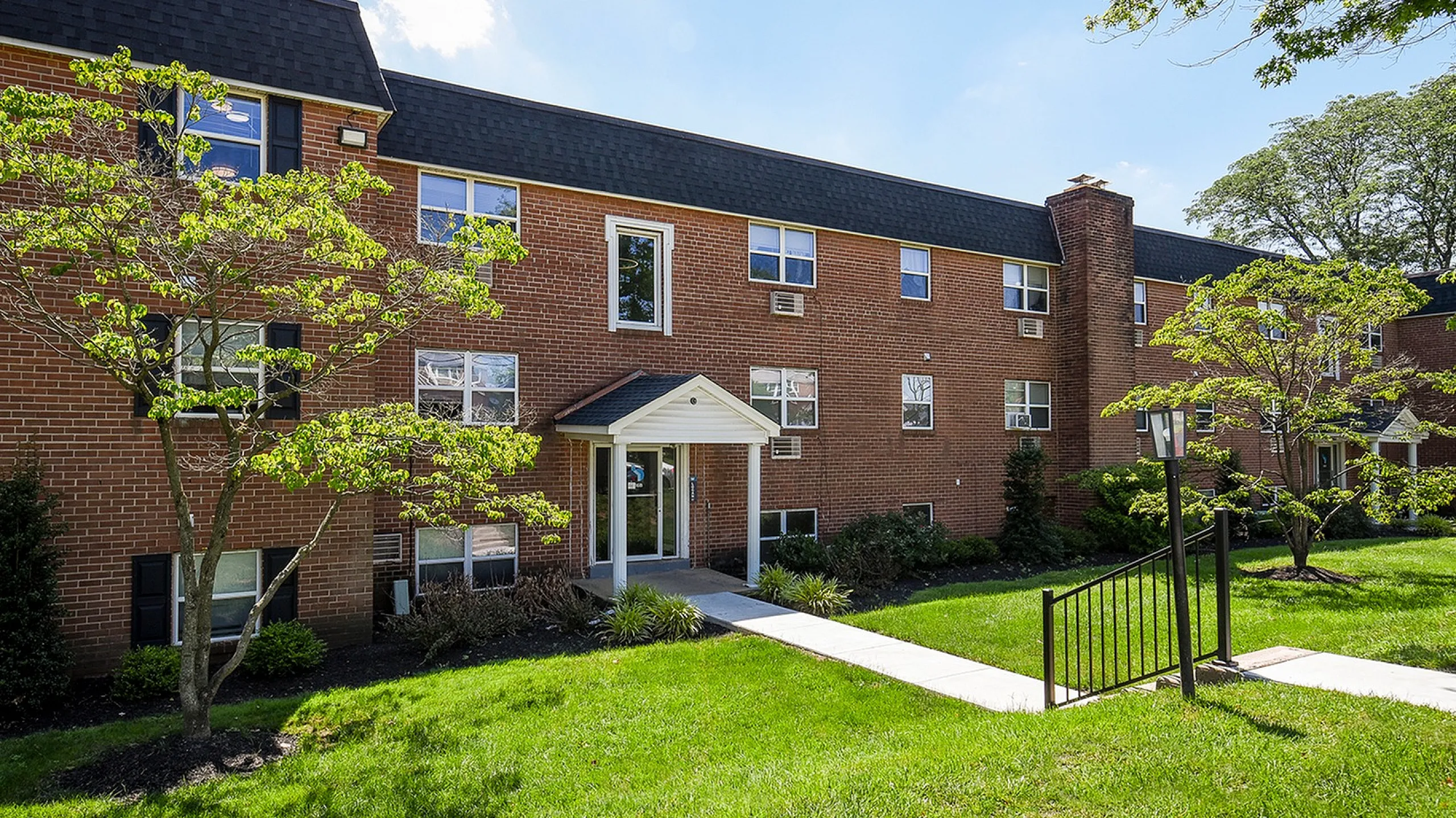 Charming brick apartment building surrounded by lush greenery and well-kept lawns, reflecting a welcoming and serene atmosphere.