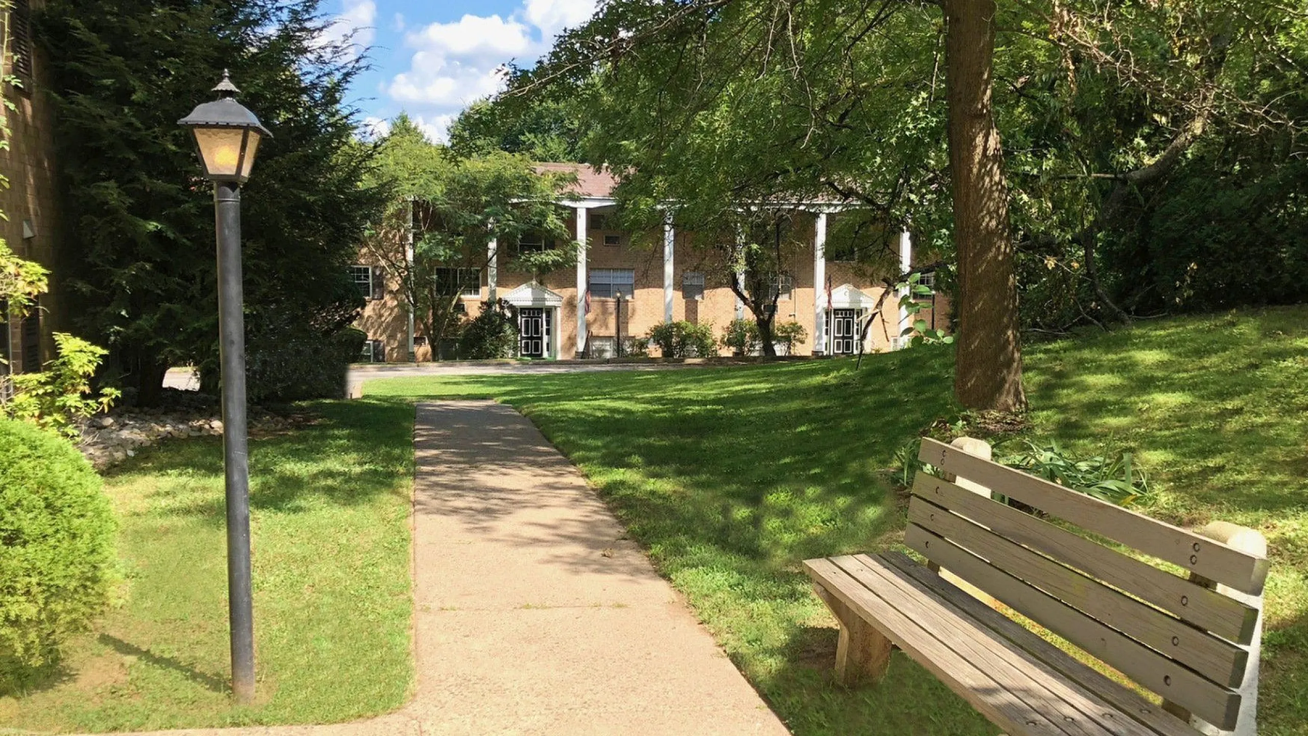 Charming apartment complex entrance with lush green lawn, shaded pathways, and inviting wooden bench.