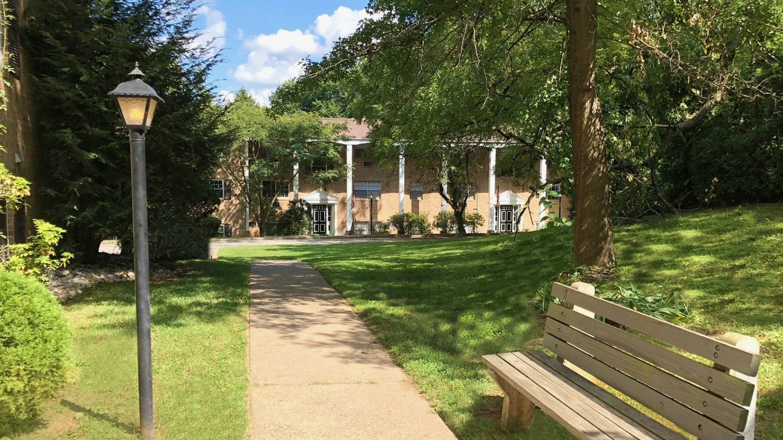 Charming apartment complex entrance with a well-maintained pathway, lush greenery, and inviting park bench, perfect for relaxation.