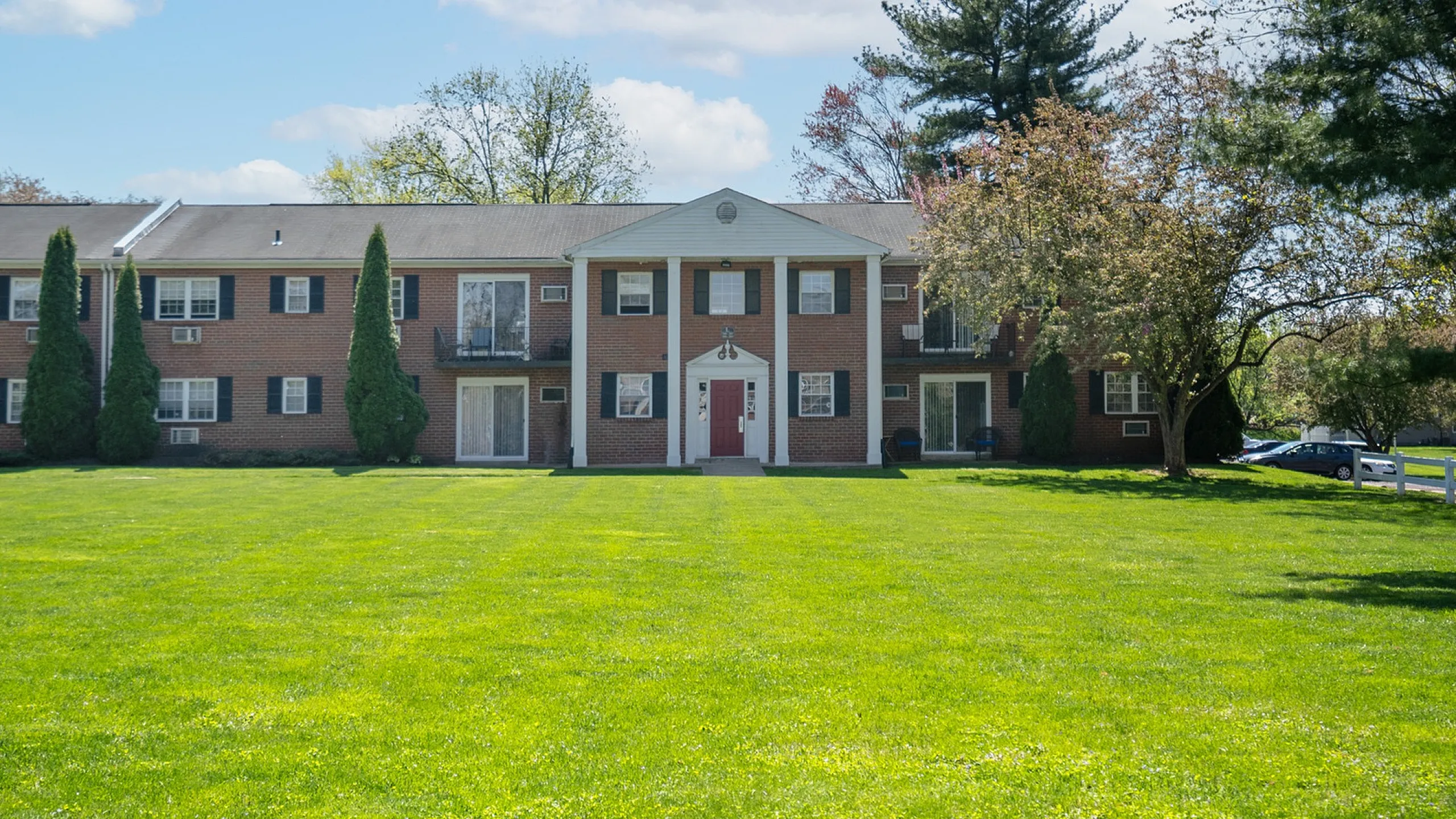Charming two-story apartment complex with classic red brick exterior, vibrant green lawn, and elegant white columns framed by lush trees.