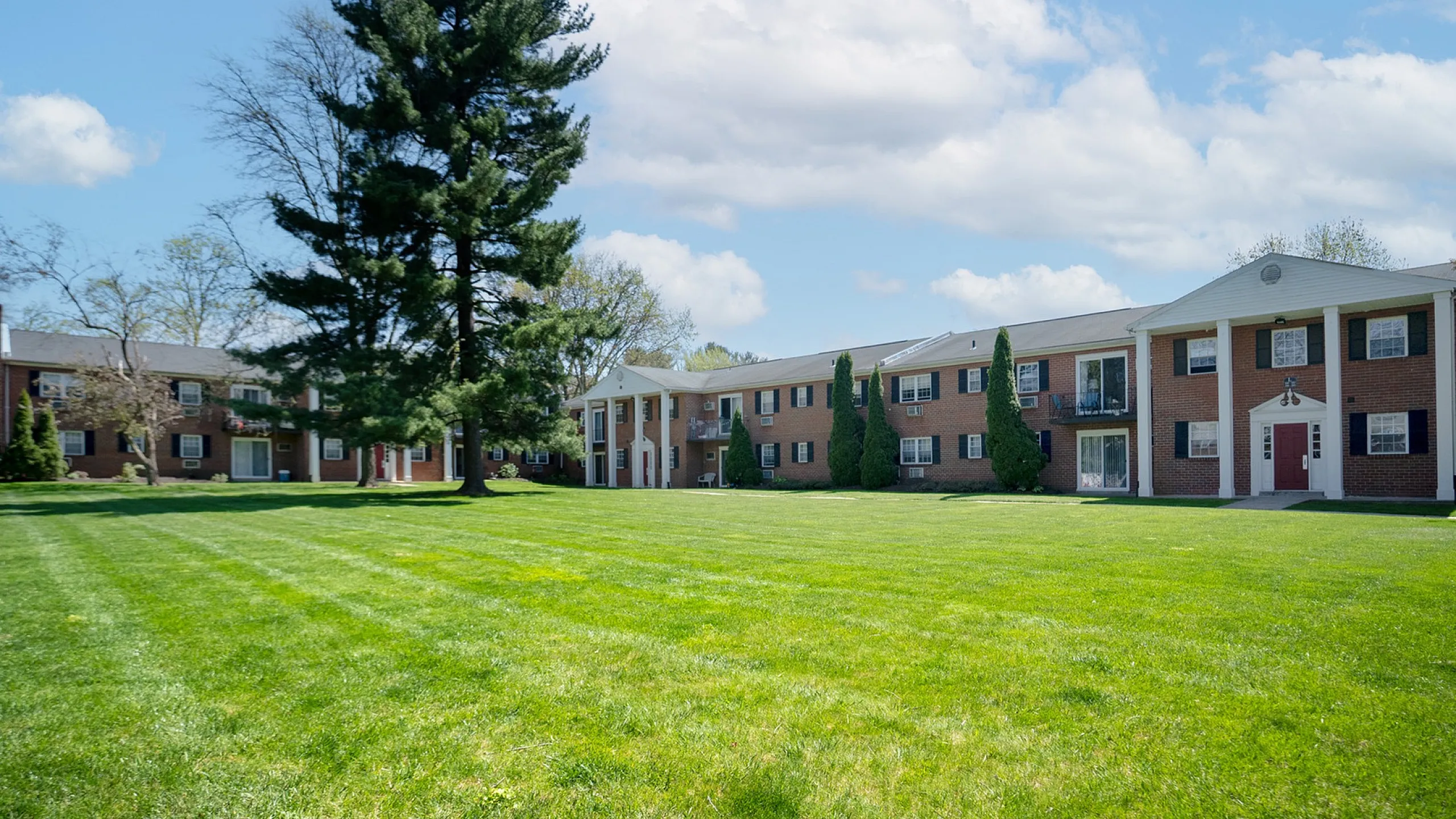 Charming apartment complex with classic brick facade, surrounded by lush green lawns and mature trees, under a bright blue sky.