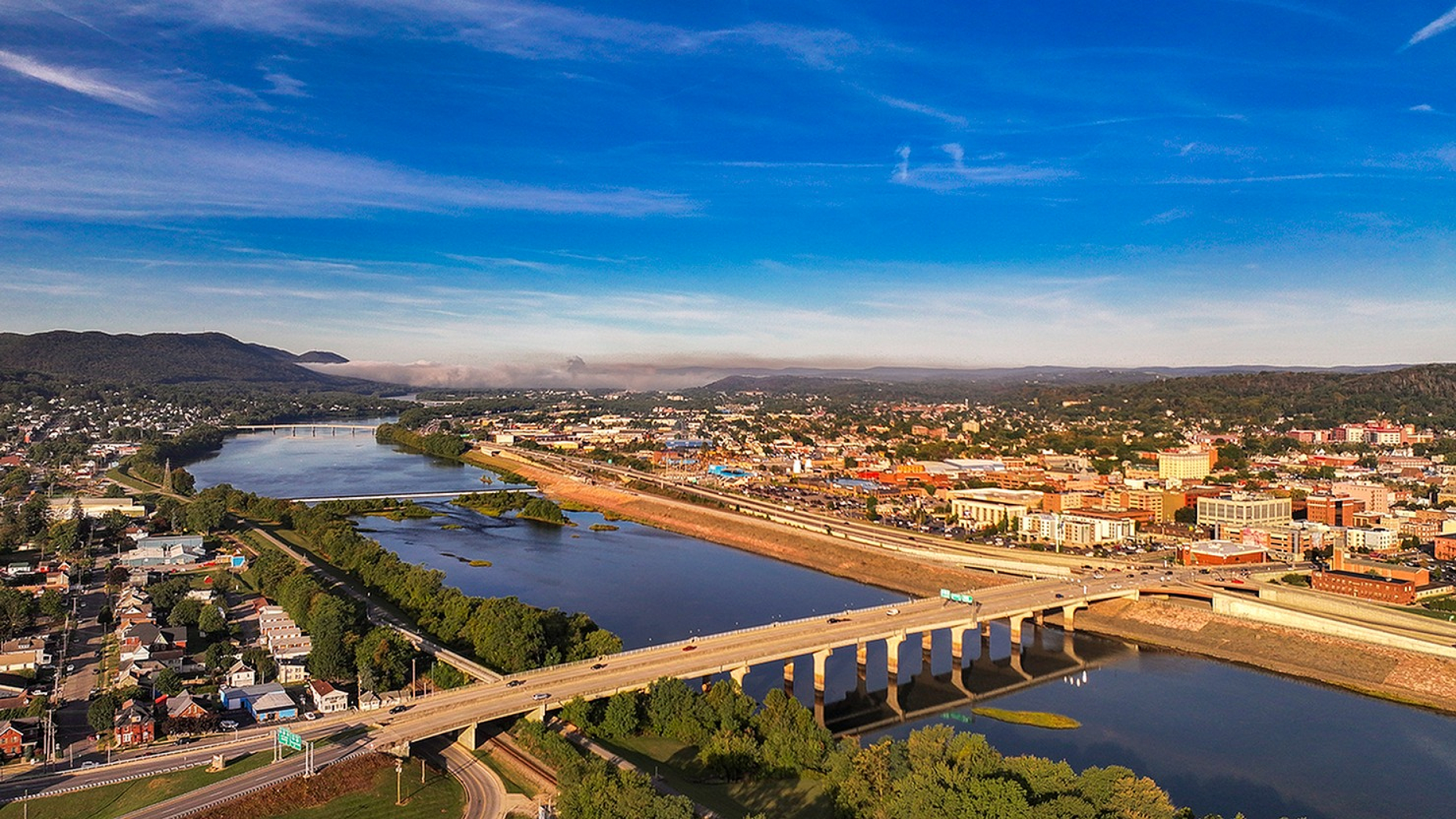 Vibrant aerial view of a scenic cityscape with a bridge spanning a calm river, surrounded by lush greenery and distant mountains under a bright blue sky.