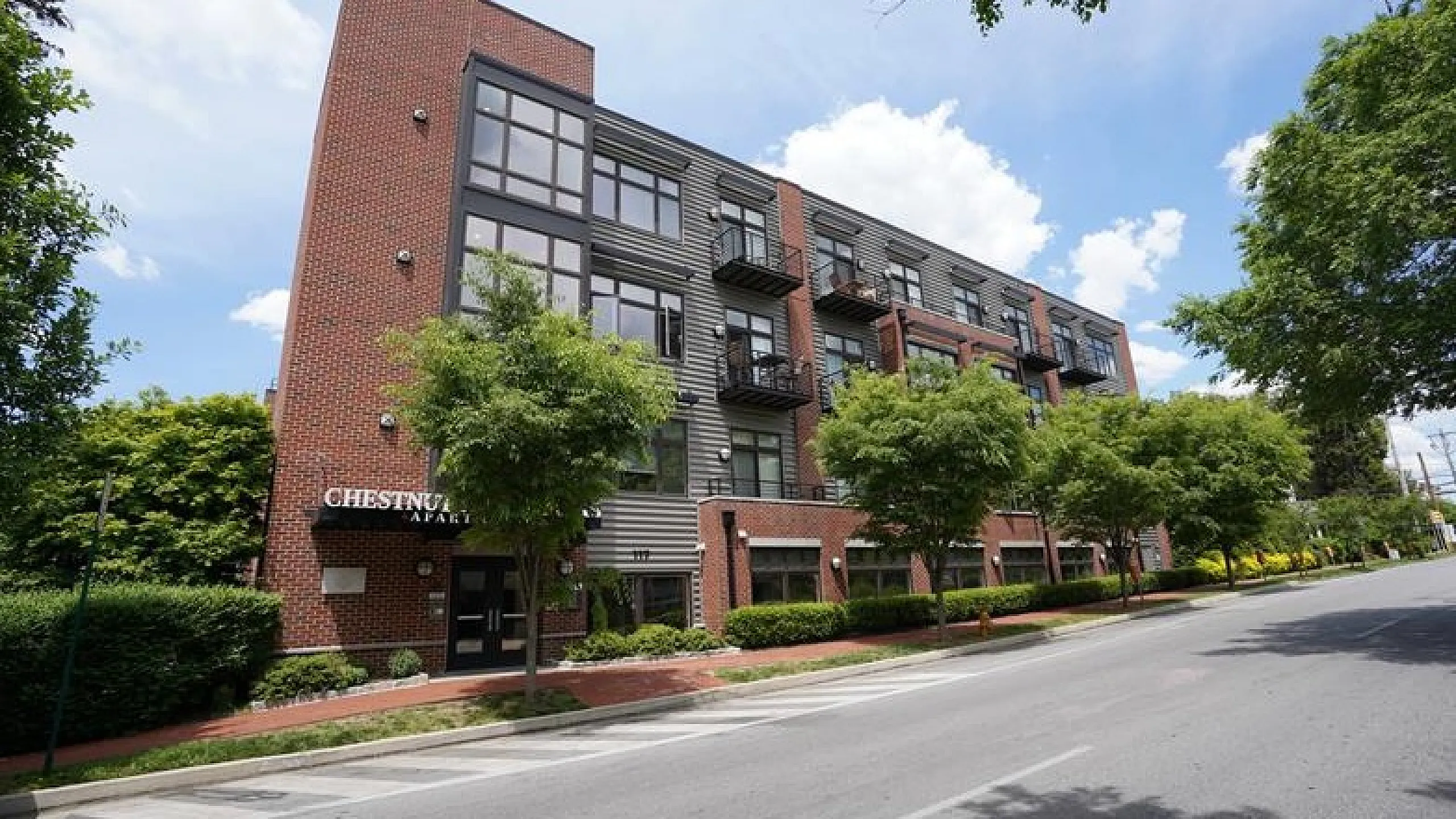Modern brick apartment building with large windows and lush trees, set on a serene street with clear blue skies.